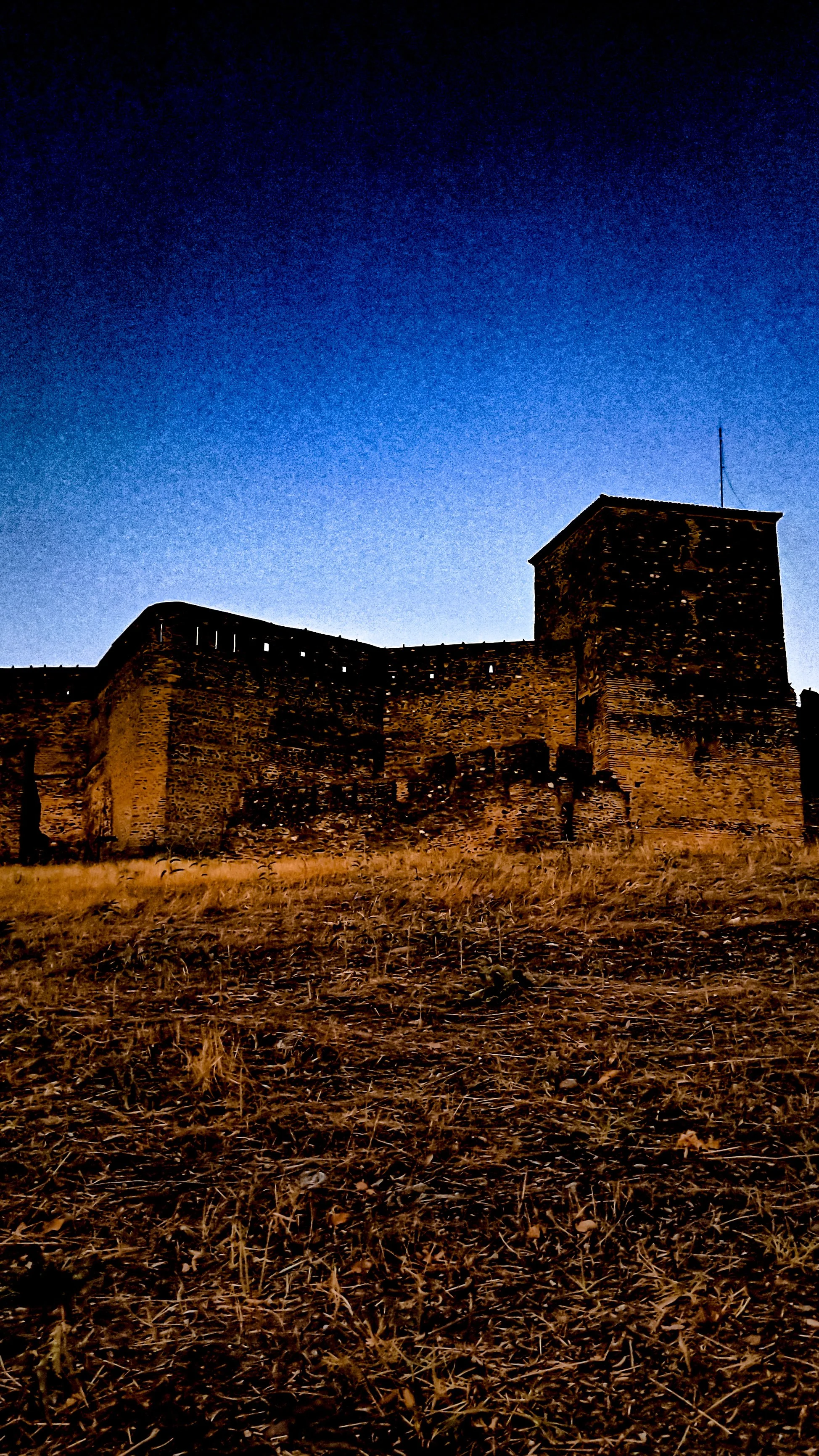 An old stone castle with high walls and a square tower, under a dark evening sky.