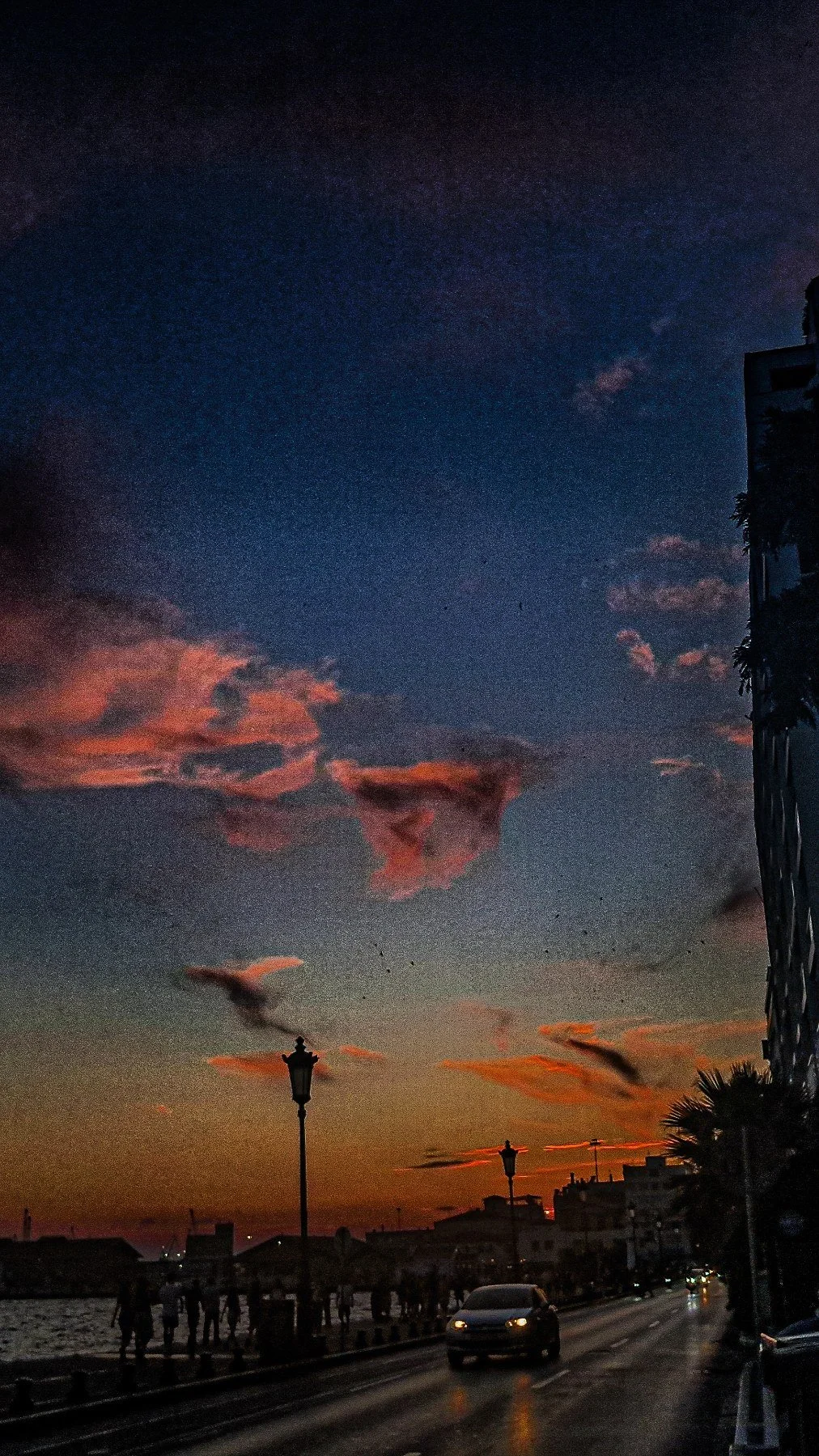 Sunset over a waterfront promenade with silhouetted people, street lamps, and buildings, and a colorful sky with pink, orange, and purple clouds.