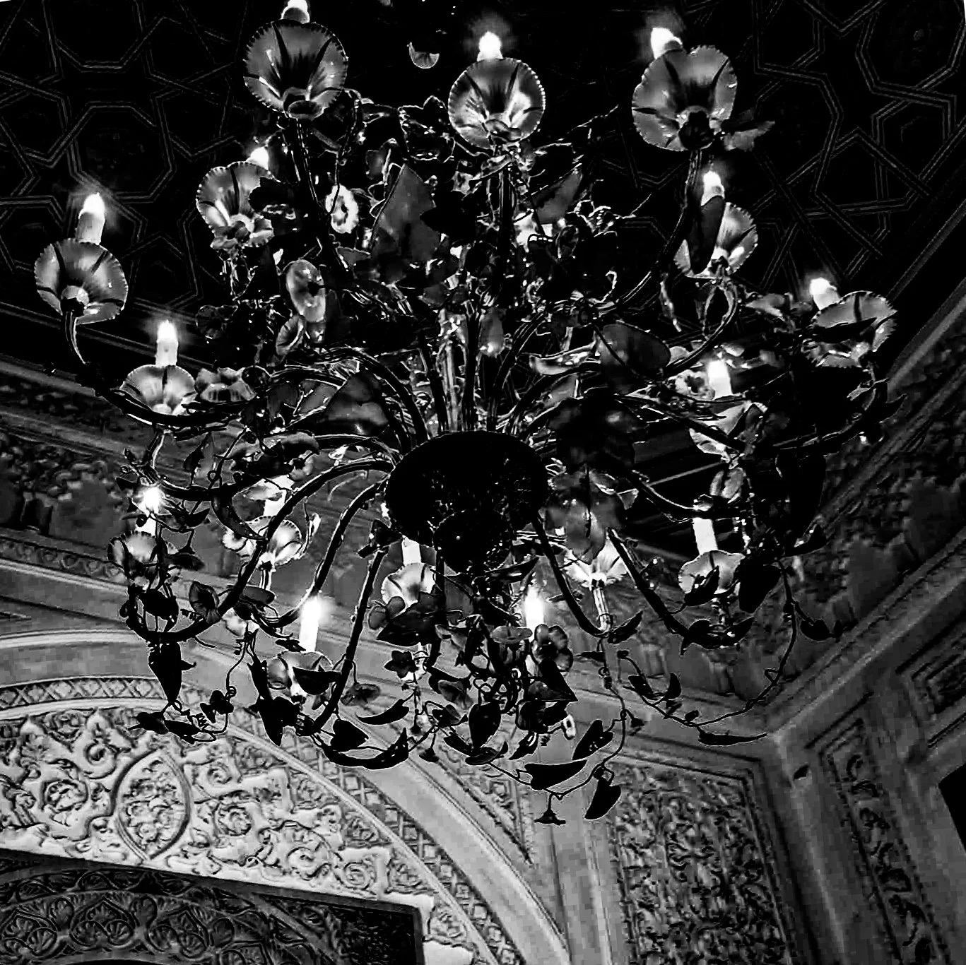 A black and white photograph of a large ornate chandelier hanging from an elaborately decorated ceiling with intricate carvings.