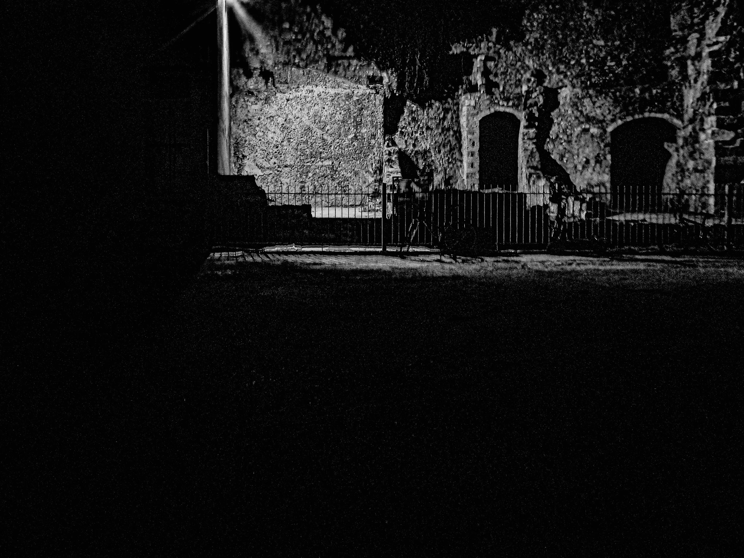 Nighttime scene of an old stone wall with three arched doorways, illuminated by moonlight or streetlight, with a fence and bicycles in the foreground.