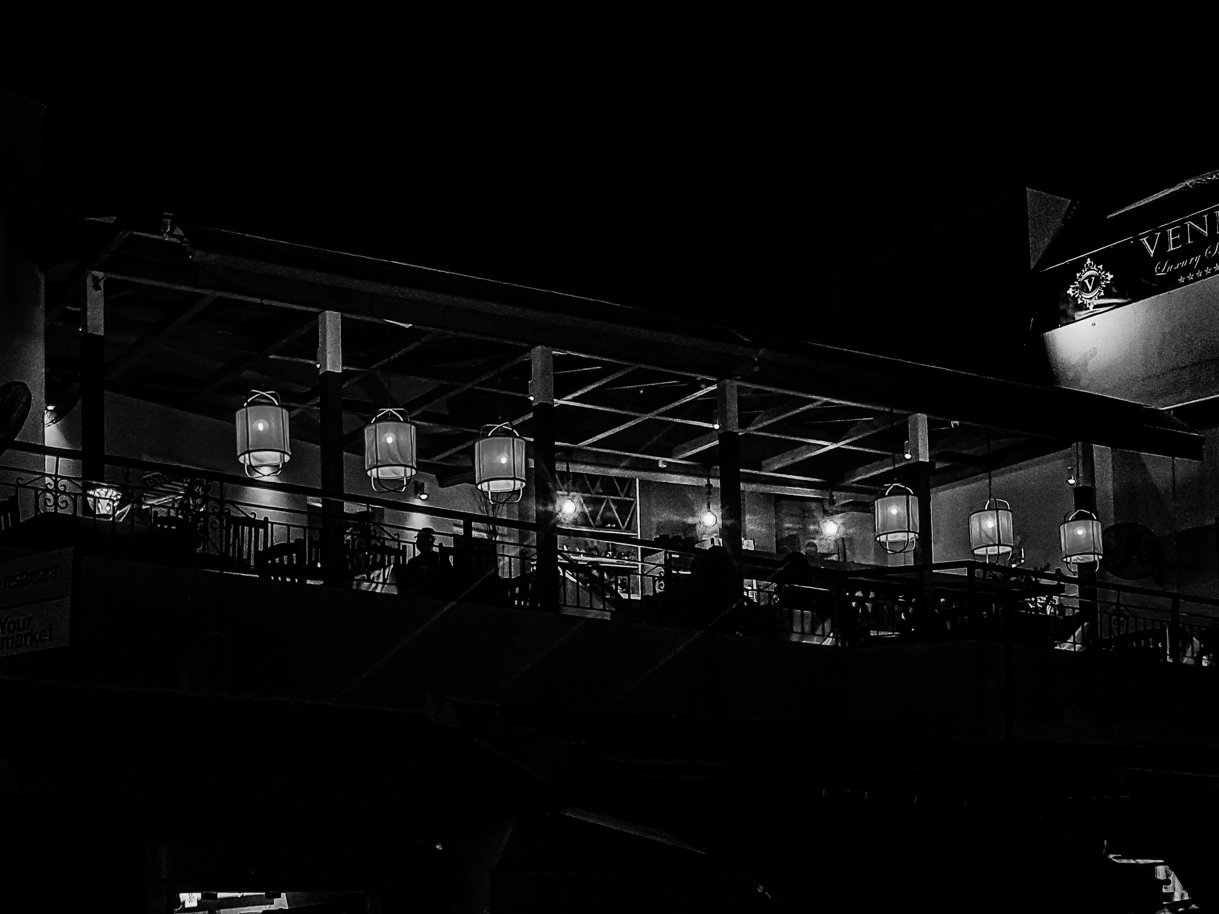 Nighttime scene inside a dimly lit restaurant or bar with hanging lantern-style lights, dark furniture, and a balcony railing, with a sign that reads 'VENA' in the background.