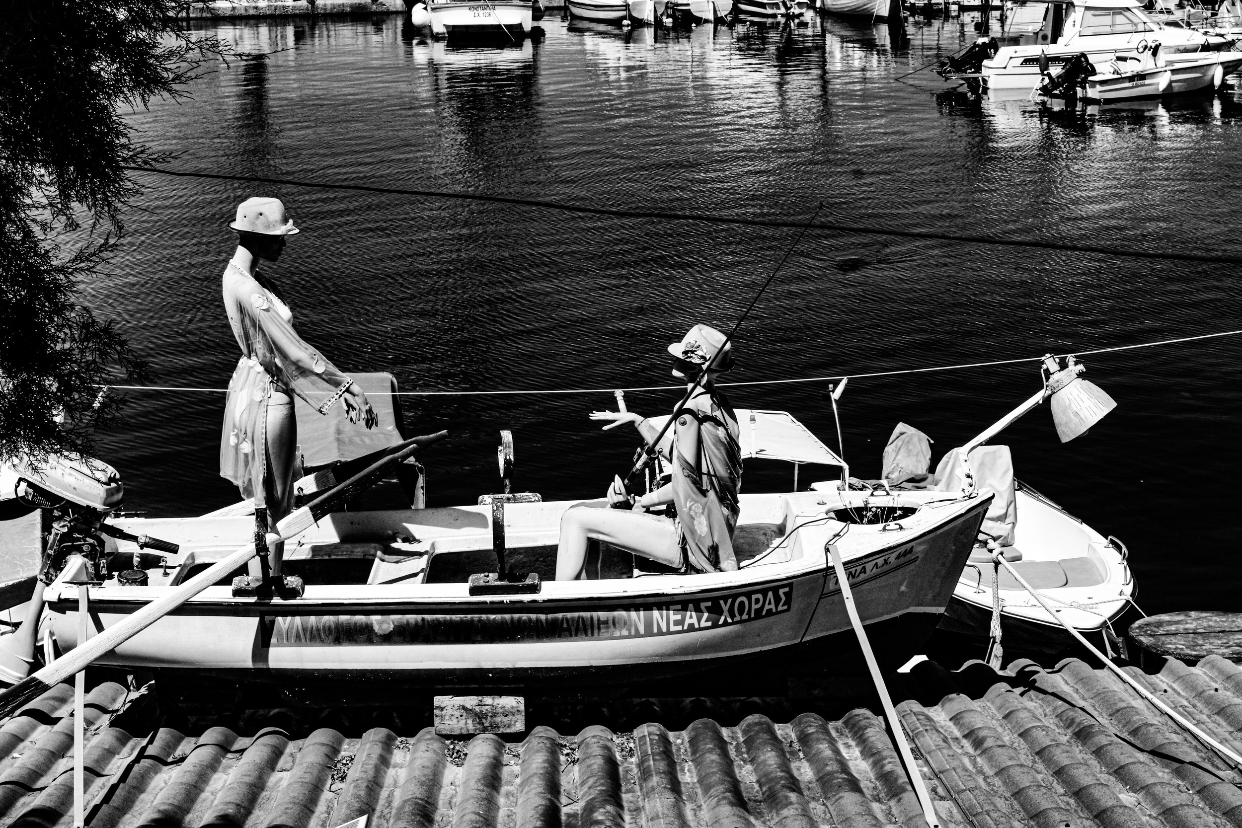 Black and white photo of a small boat docked near the shore, with two children wearing hats playing inside it, and a woman standing nearby. Several other boats are visible in the water, and a tree branches is partially blocking the view on the left.