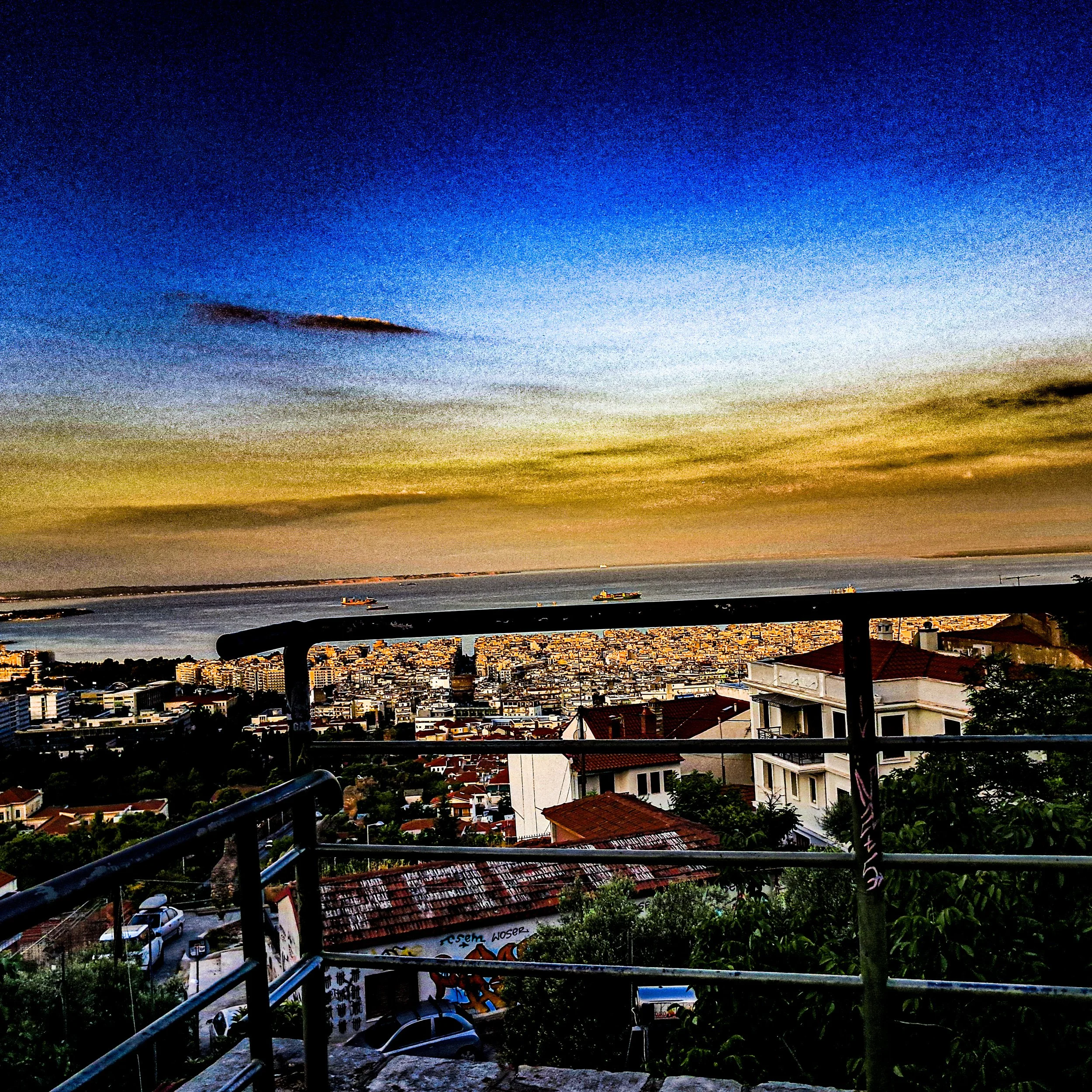 Cityscape view during sunset from a balcony with railings, showing buildings, a body of water, and boats in the distance under a colorful sky.