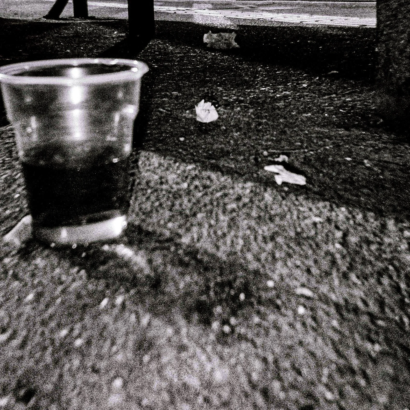 Black and white image depicting a clear glass of water, a crumpled piece of paper, and a small white object on a textured surface, with a partial view of a wooden object in the background.