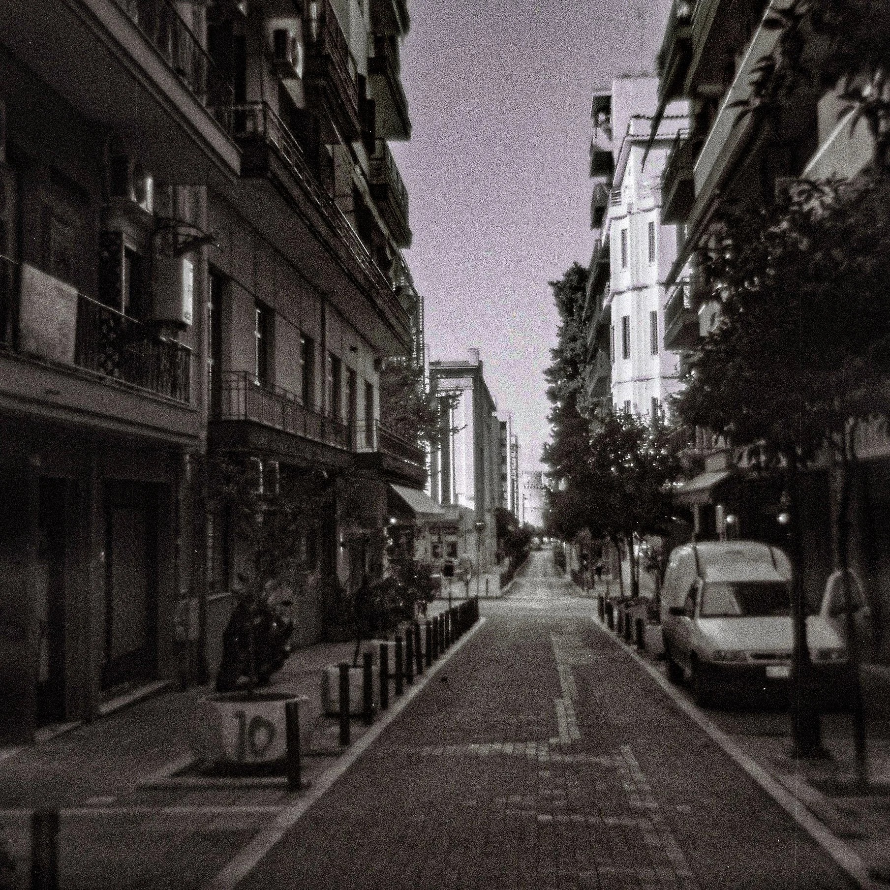 Nighttime scene of a narrow city street with parked cars on both sides, tall apartment buildings with balconies and air conditioning units, and trees lining the sidewalk.