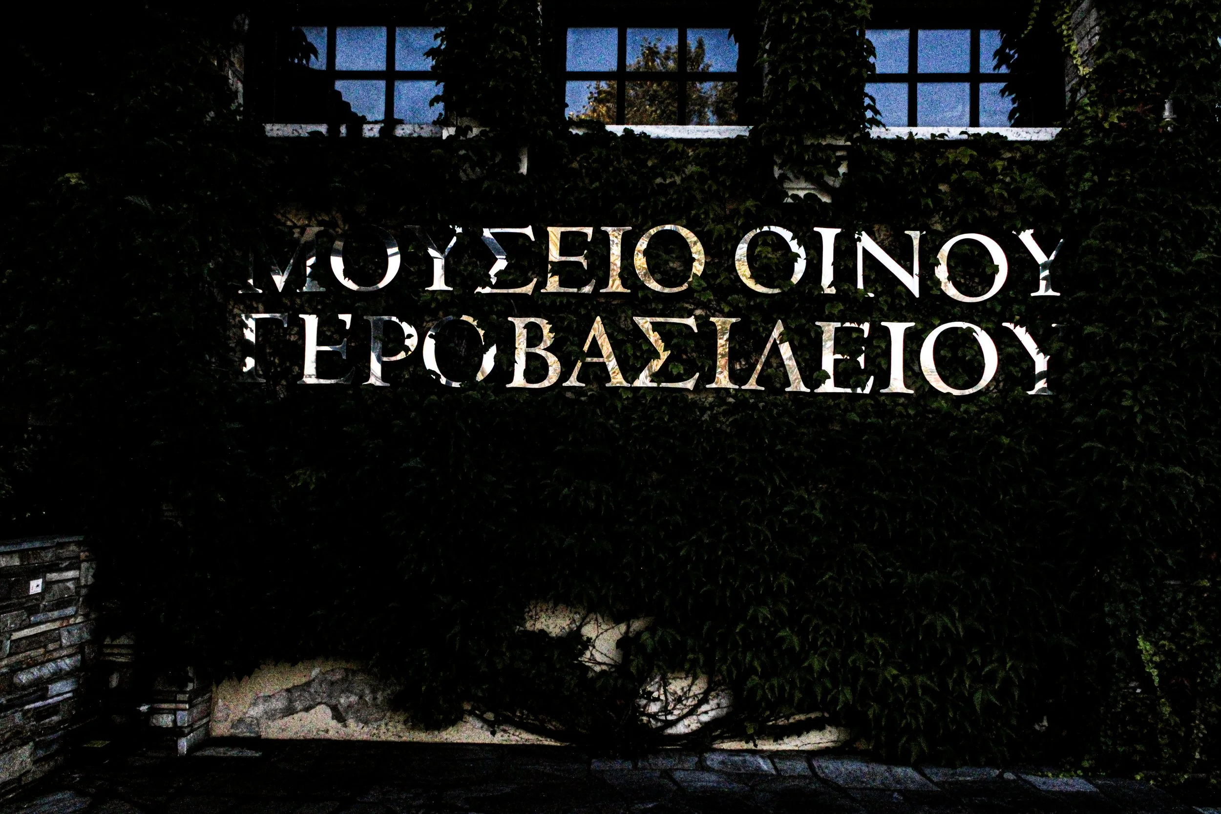 A dark outdoor scene featuring illuminated metal letters spelling 'Museum of Contemporary Epobasileio' against a background of green ivy-covered wall and a window with a view of trees and a blue sky.