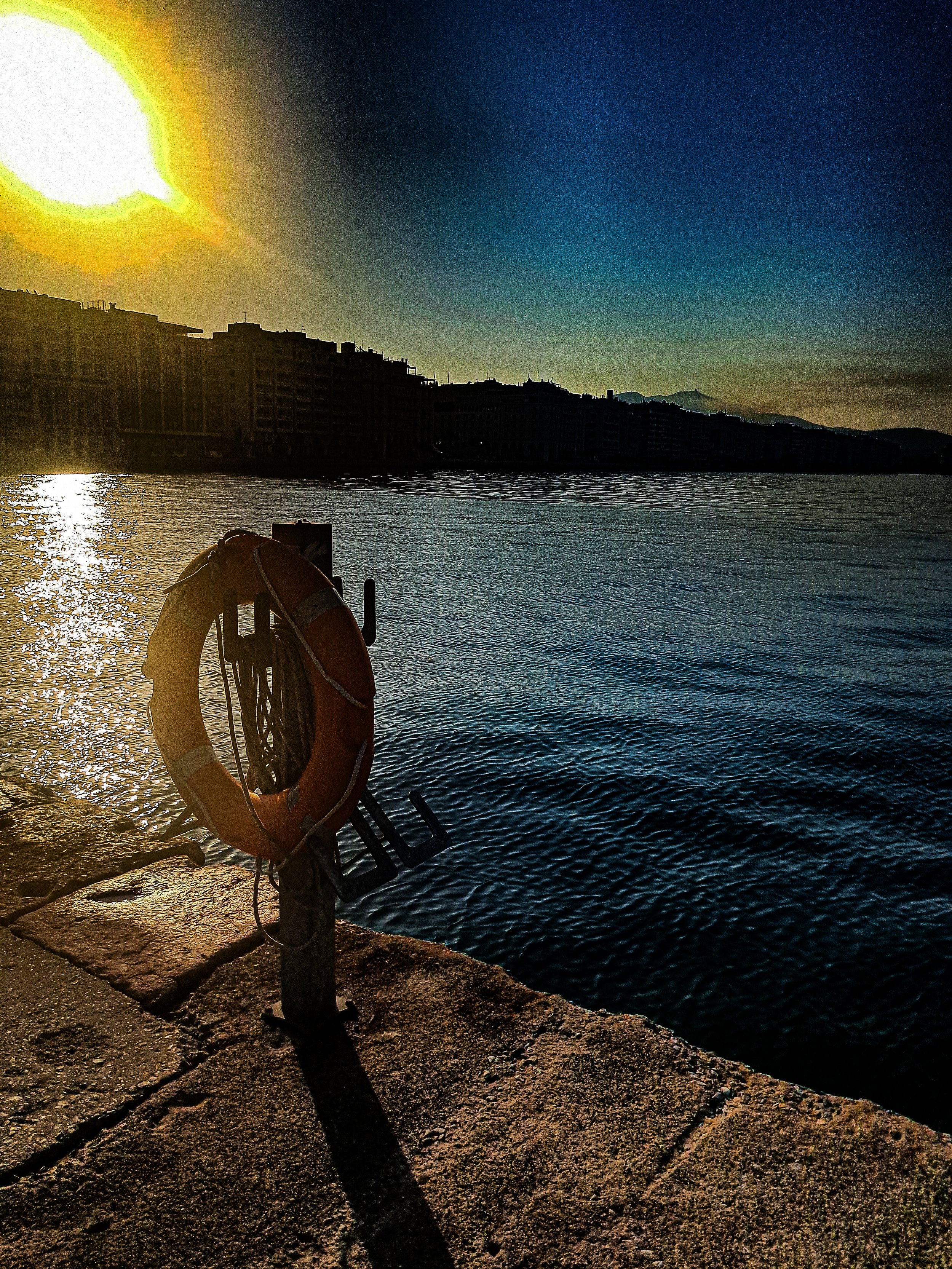 Sunset over a body of water with buildings in the background and a life preserver hanging on a post in the foreground.