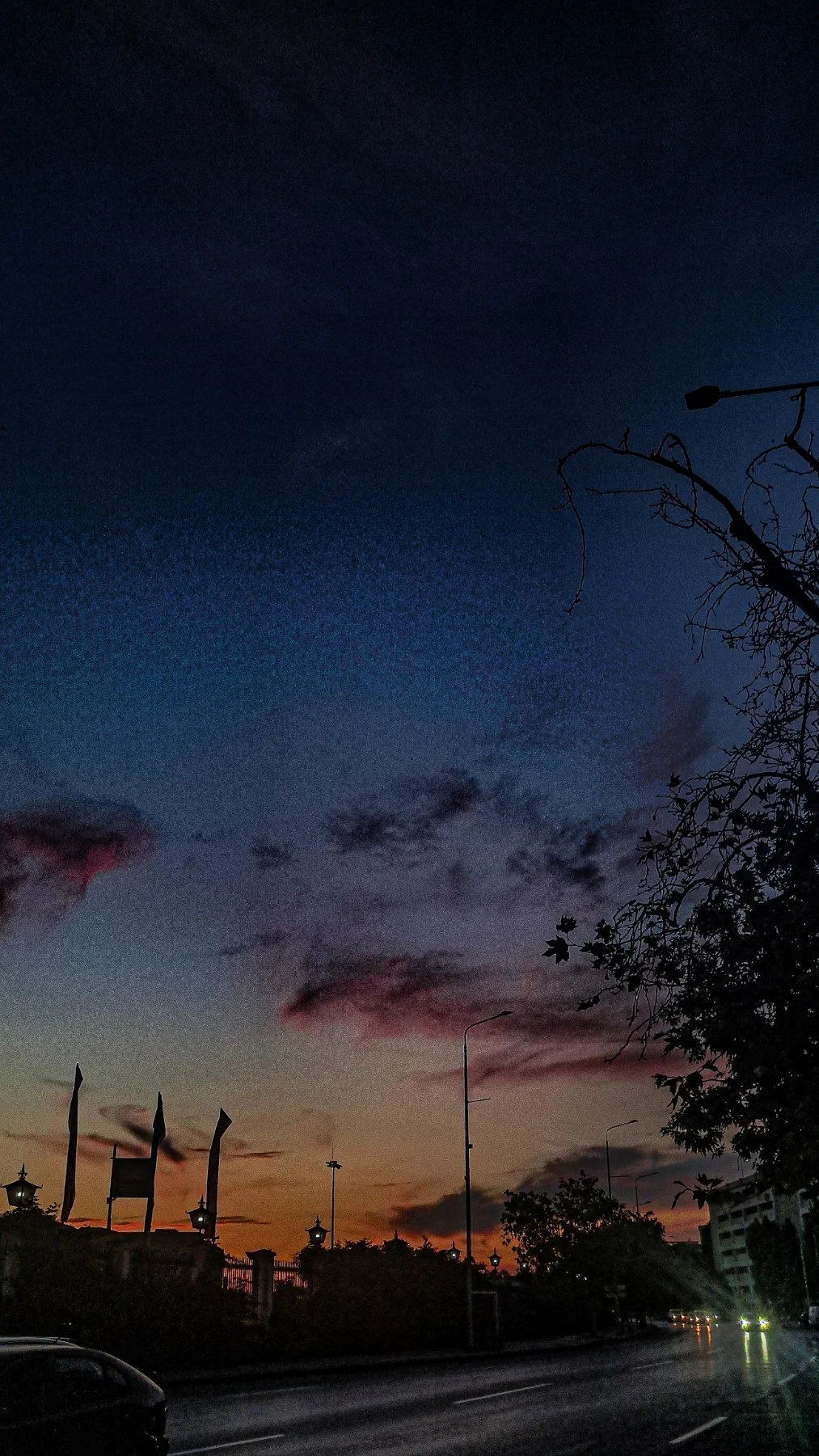 Darkening sky during sunset with clouds, silhouettes of leafless trees and street lamps along a road, some cars with headlights on in a cityscape.