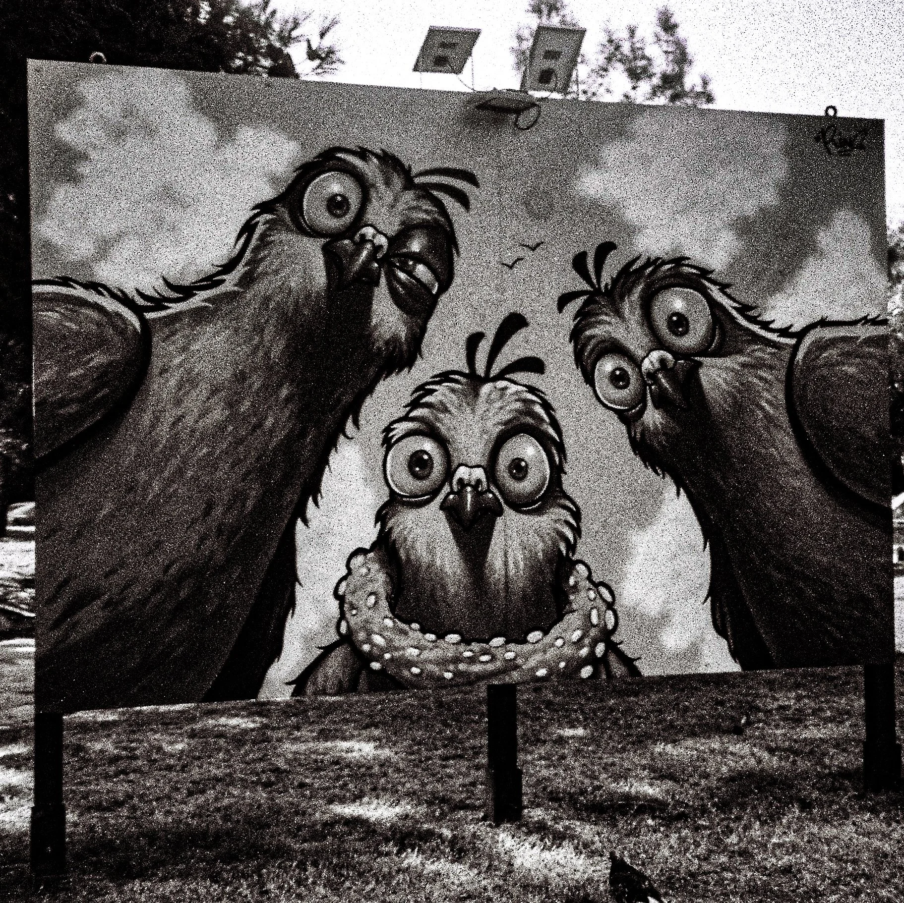 Black and white mural depicting three cartoon chickens with large eyes, combs, and beaks, standing side by side in front of a cloudy sky background with a few birds flying.