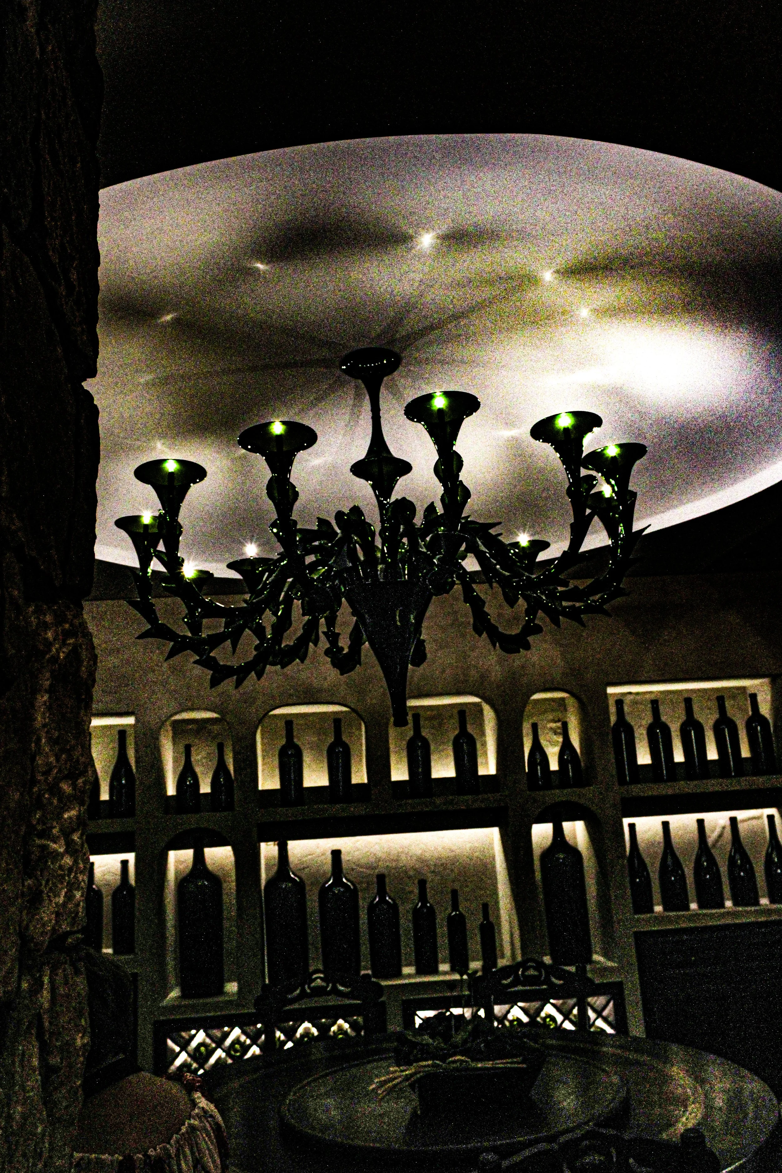 A decorative black chandelier with lit green lights hanging from a textured ceiling in a room with wine bottle shelves.