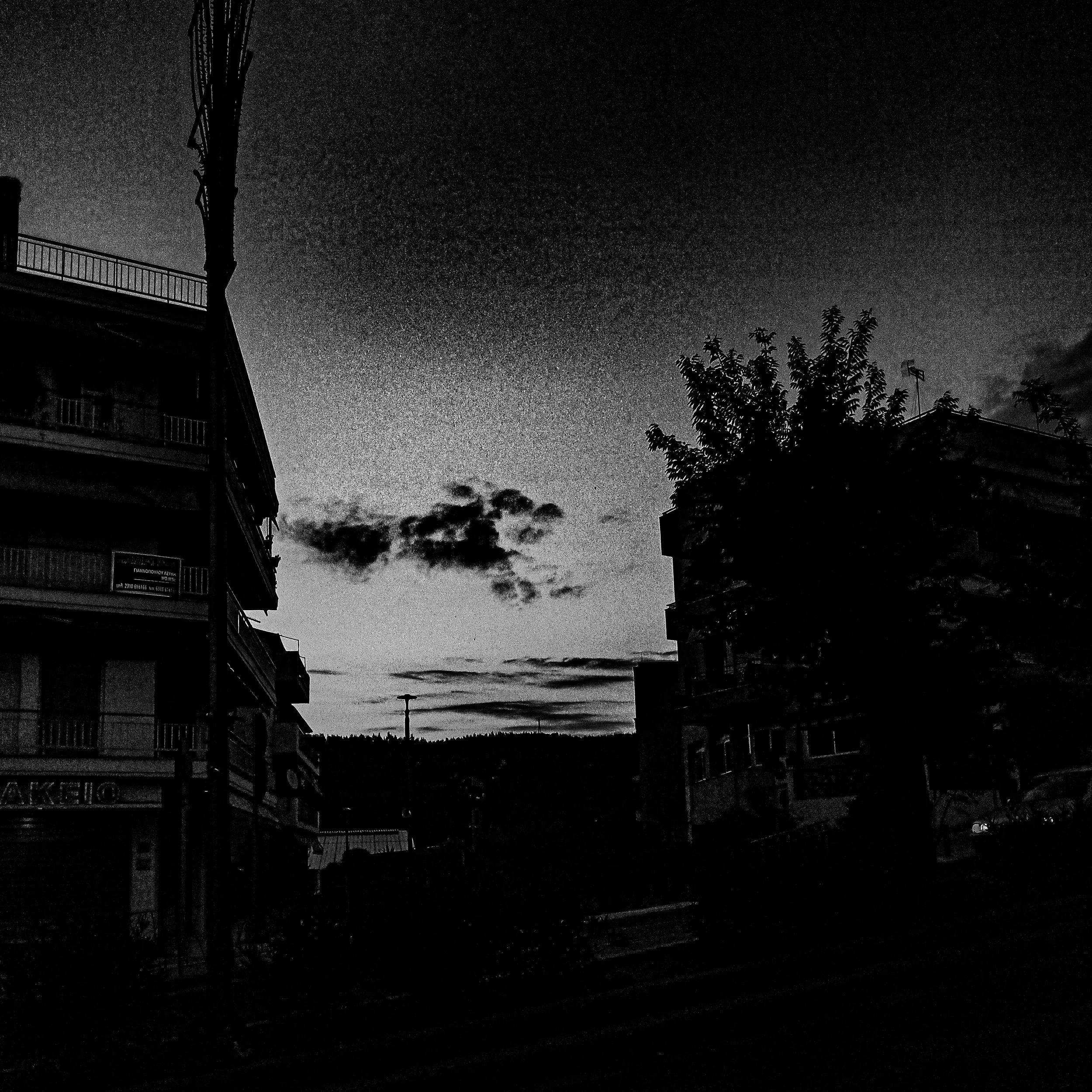 A black and white photo of a city street at night, with buildings on the left, a tree on the right, and a cloudy sky overhead.