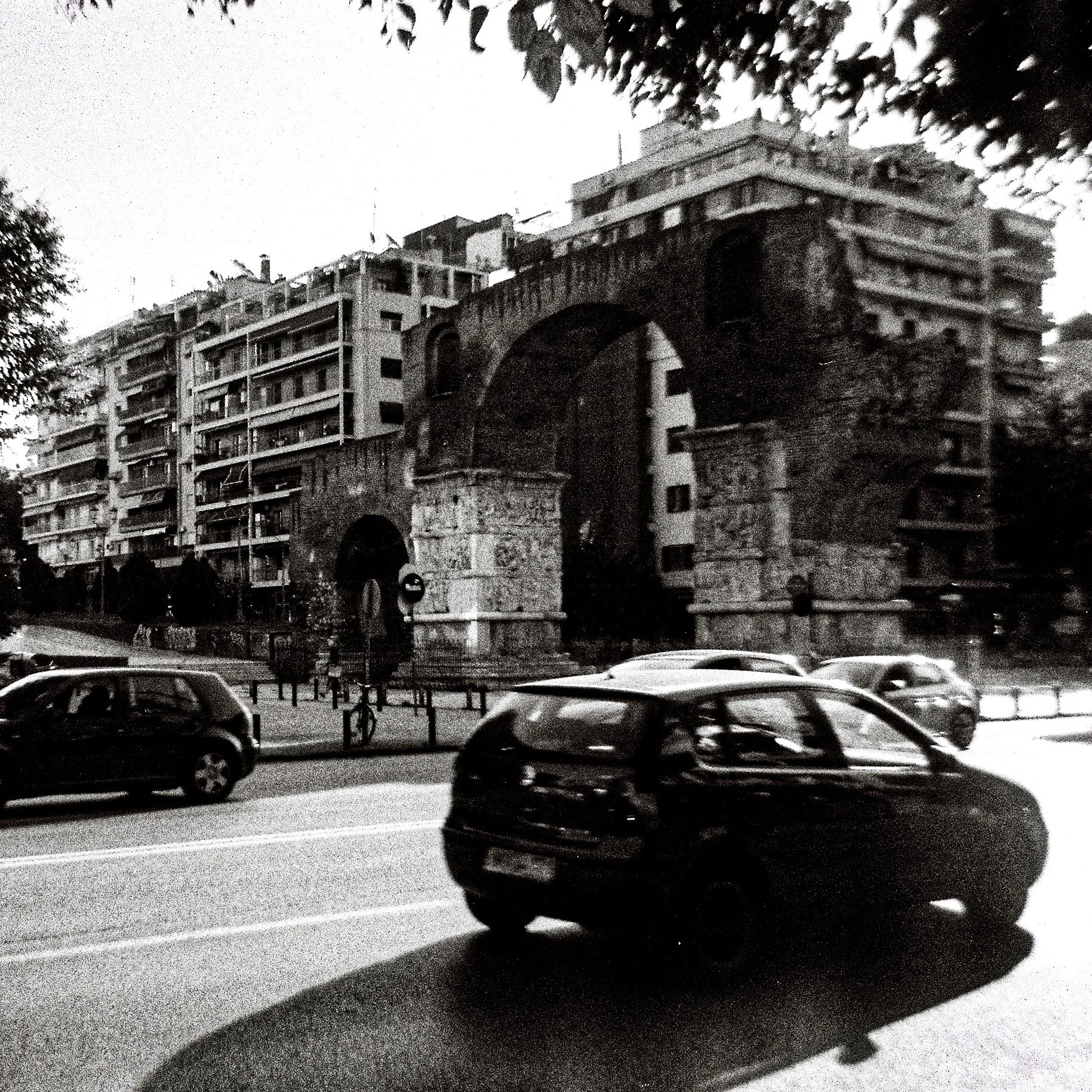 Black and white photo of a large stone archway and a modern multi-story building in the background, viewed from a street with parked cars and trees.