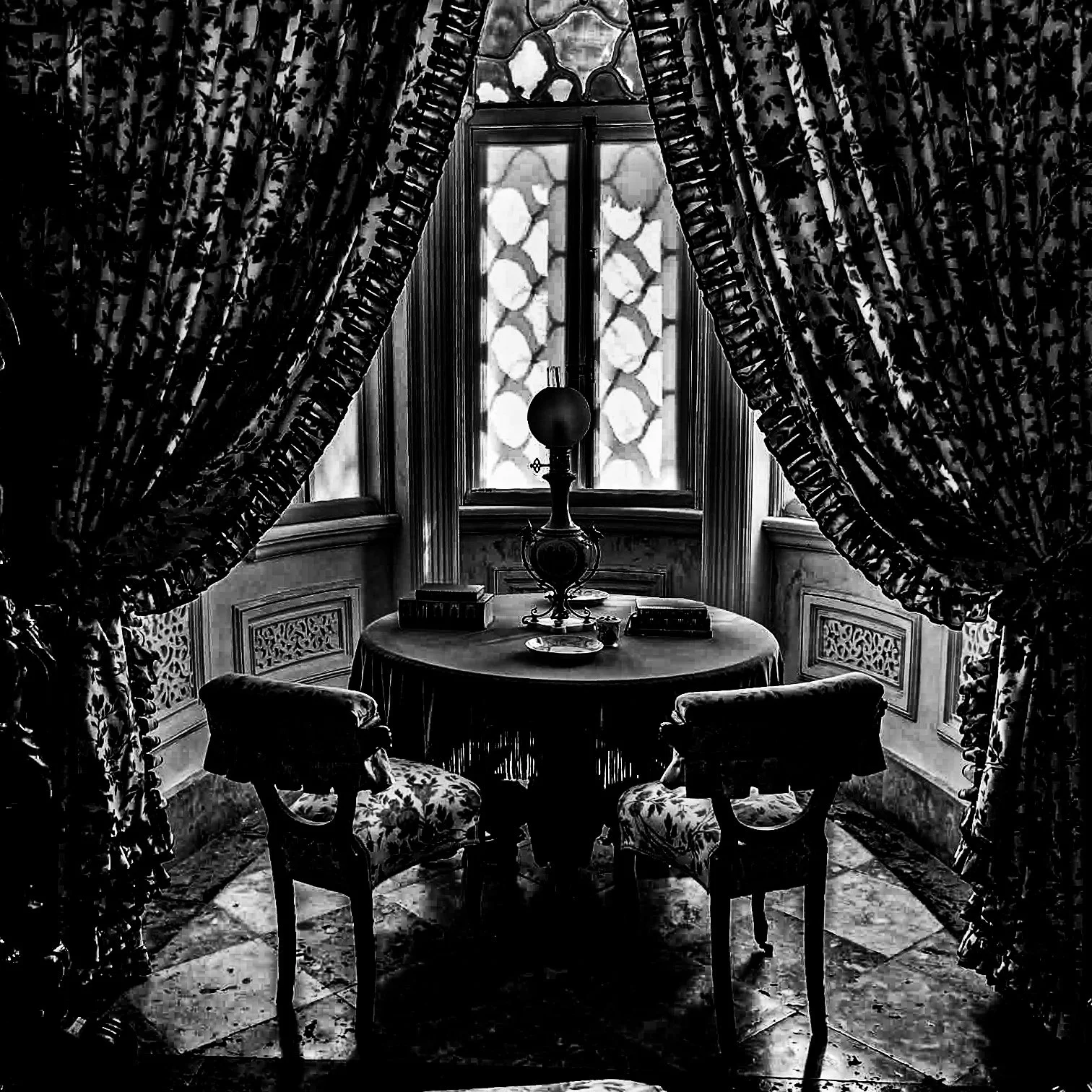 A vintage-style interior with a round table covered by a cloth, surrounded by four chairs with floral upholstery, and a central decorative lamp on the table. Behind the table is a large window with patterned glass, flanked by ornate curtains. The roo