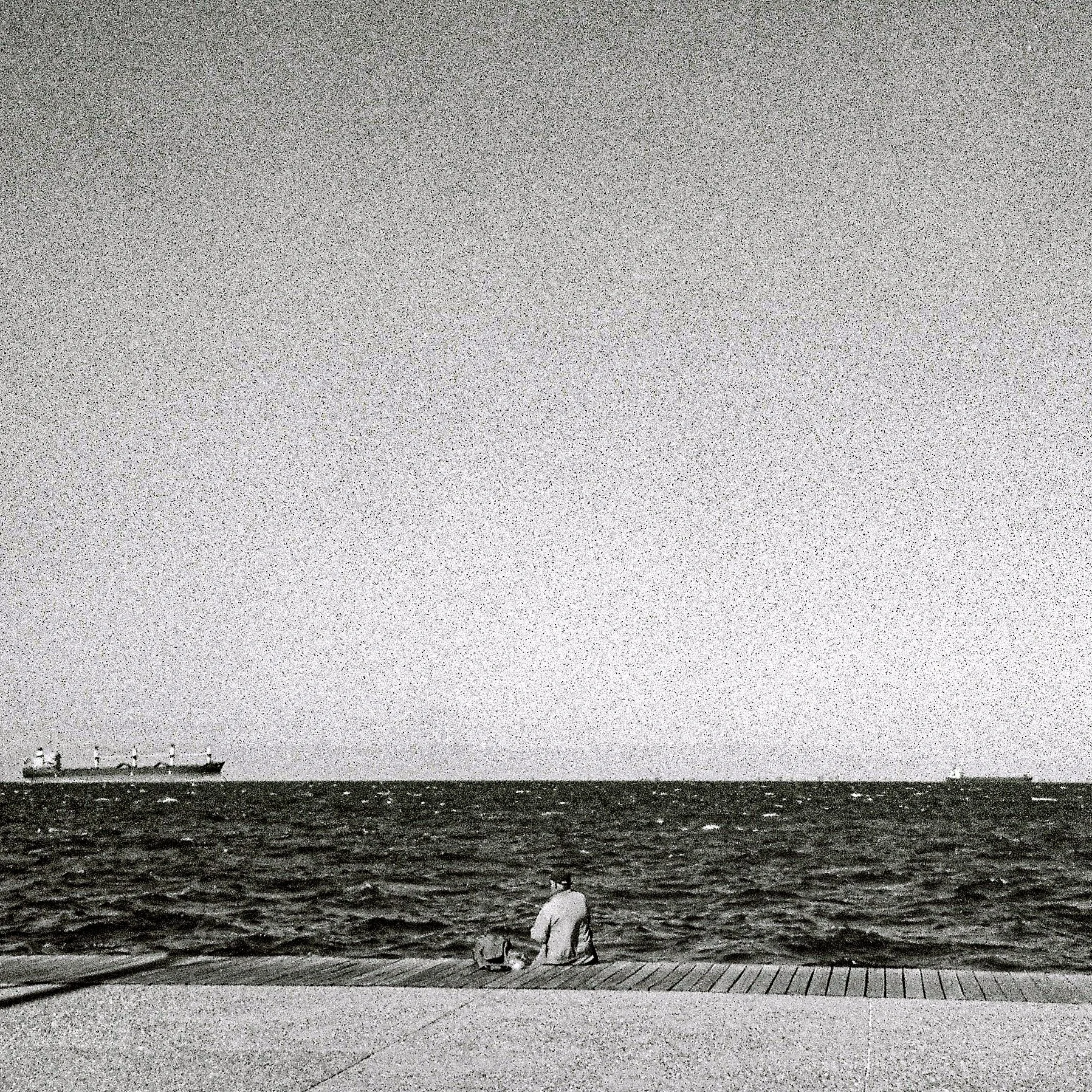 A woman sitting on a wooden walkway at the edge of the water, looking at a large cruise ship traveling across the ocean under a cloudy sky.