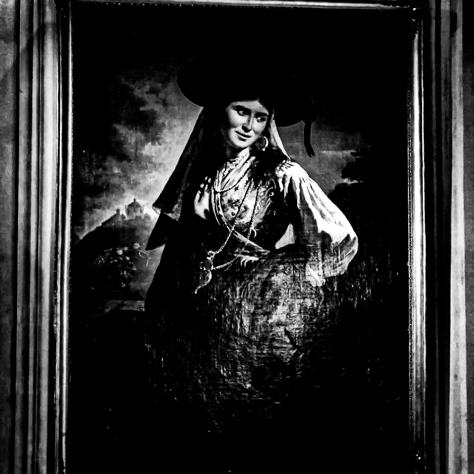A black and white portrait of a woman in traditional Mexican attire, wearing a wide sombrero, earrings, and layered necklaces, with a hand on her hip and a slight smile, inside a frame.