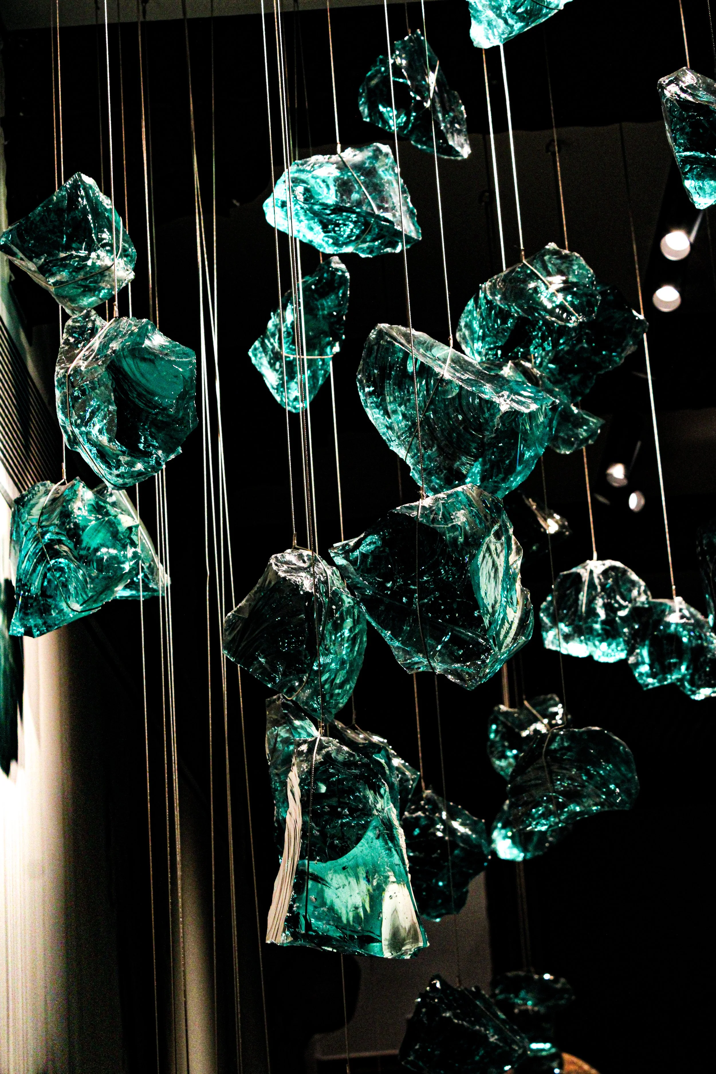 Glass art installation with irregular shaped, teal-colored glass pieces suspended from thin wires against a dark background.