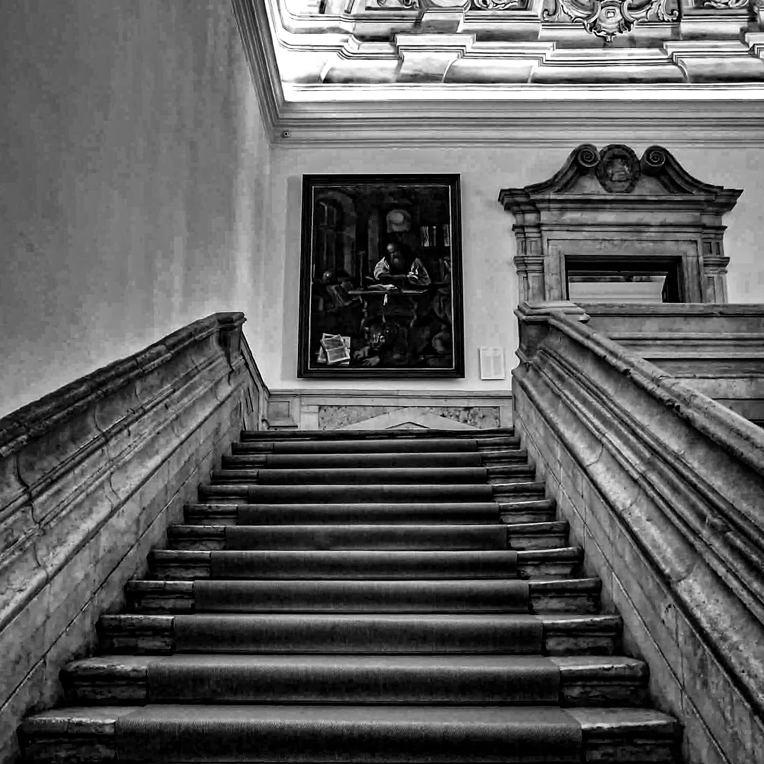 Black and white photo of a staircase with ornate wooden railing, leading up to a wall decorated with a large framed painting of a man working at a desk surrounded by books, with a cupboard and skulls visible in the background.