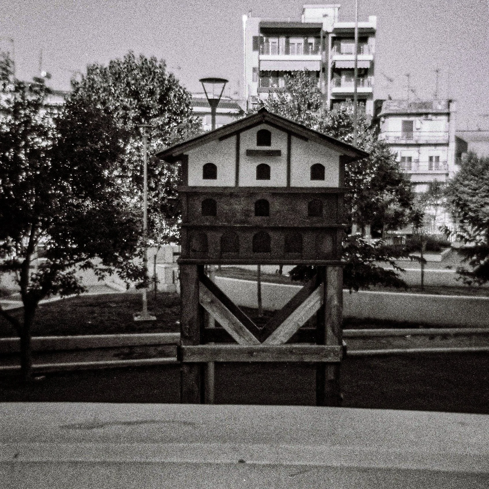 A black and white photo of a small wooden birdhouse on a stand, with residential buildings in the background and trees around.