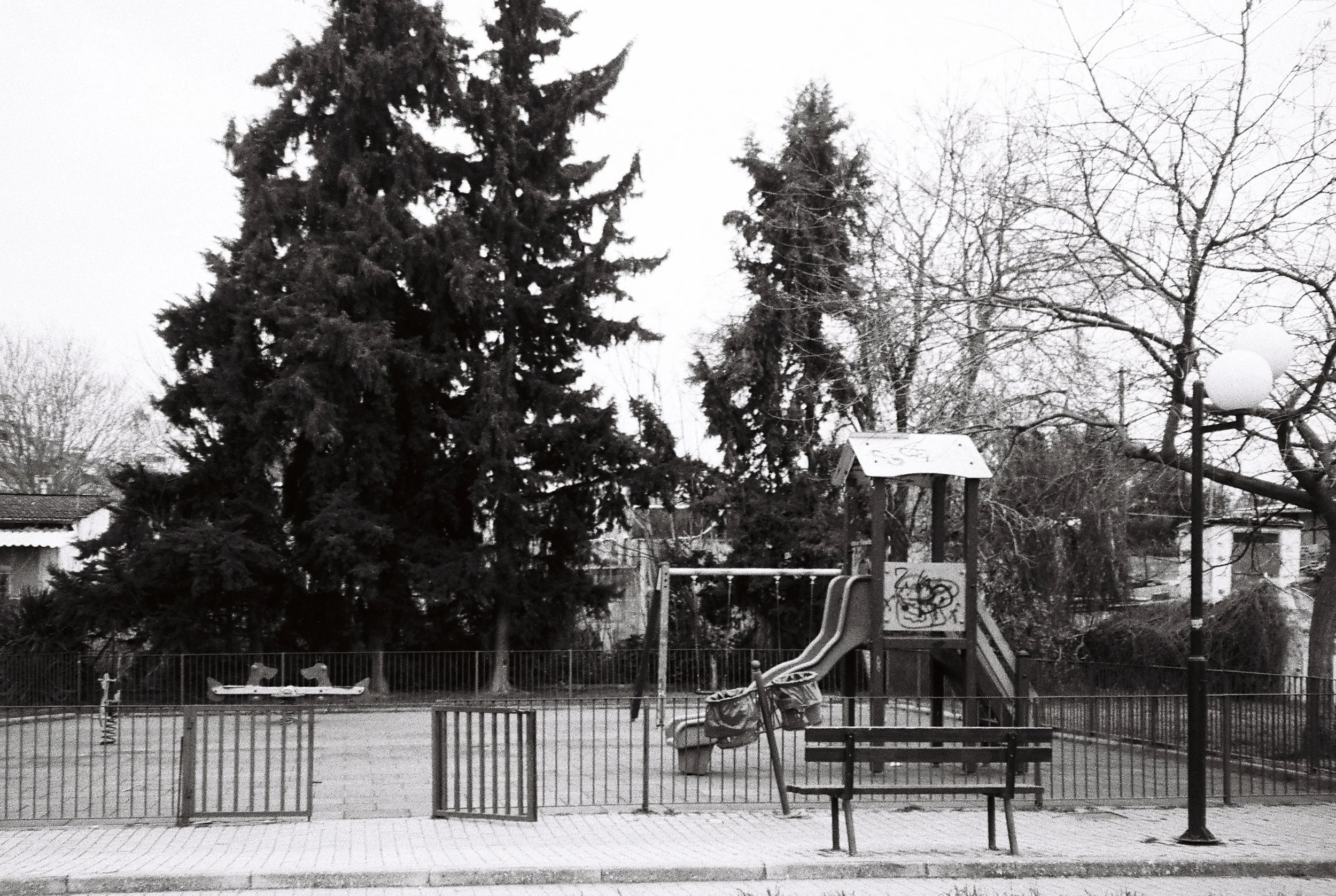 A playground area with a slide, swings, and a fenced border, located near trees with lack of leaves, suggesting winter, with a cloudy sky overhead.