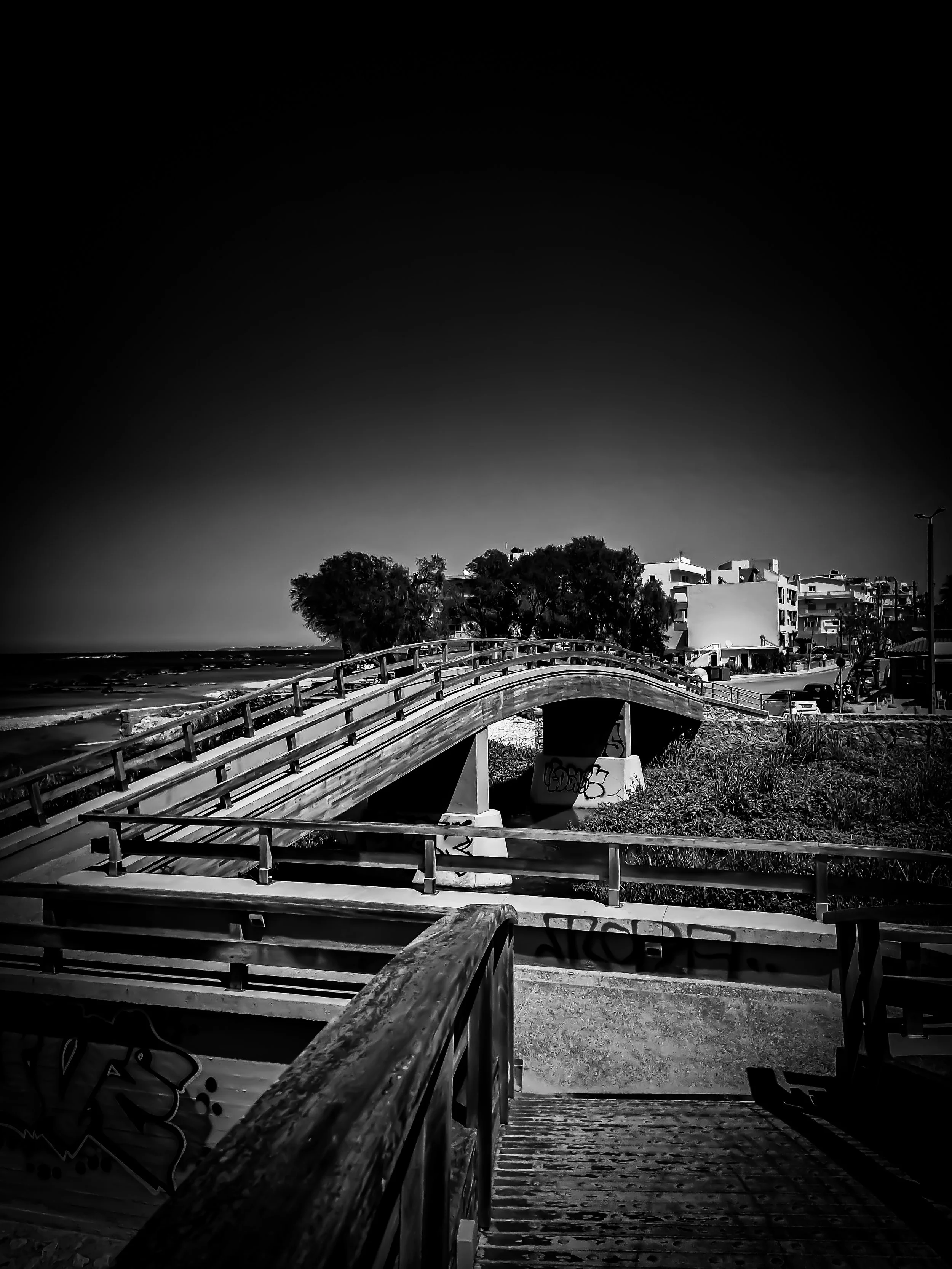 Black and white photo of a wooden bridge over a small creek with trees and buildings in the background.