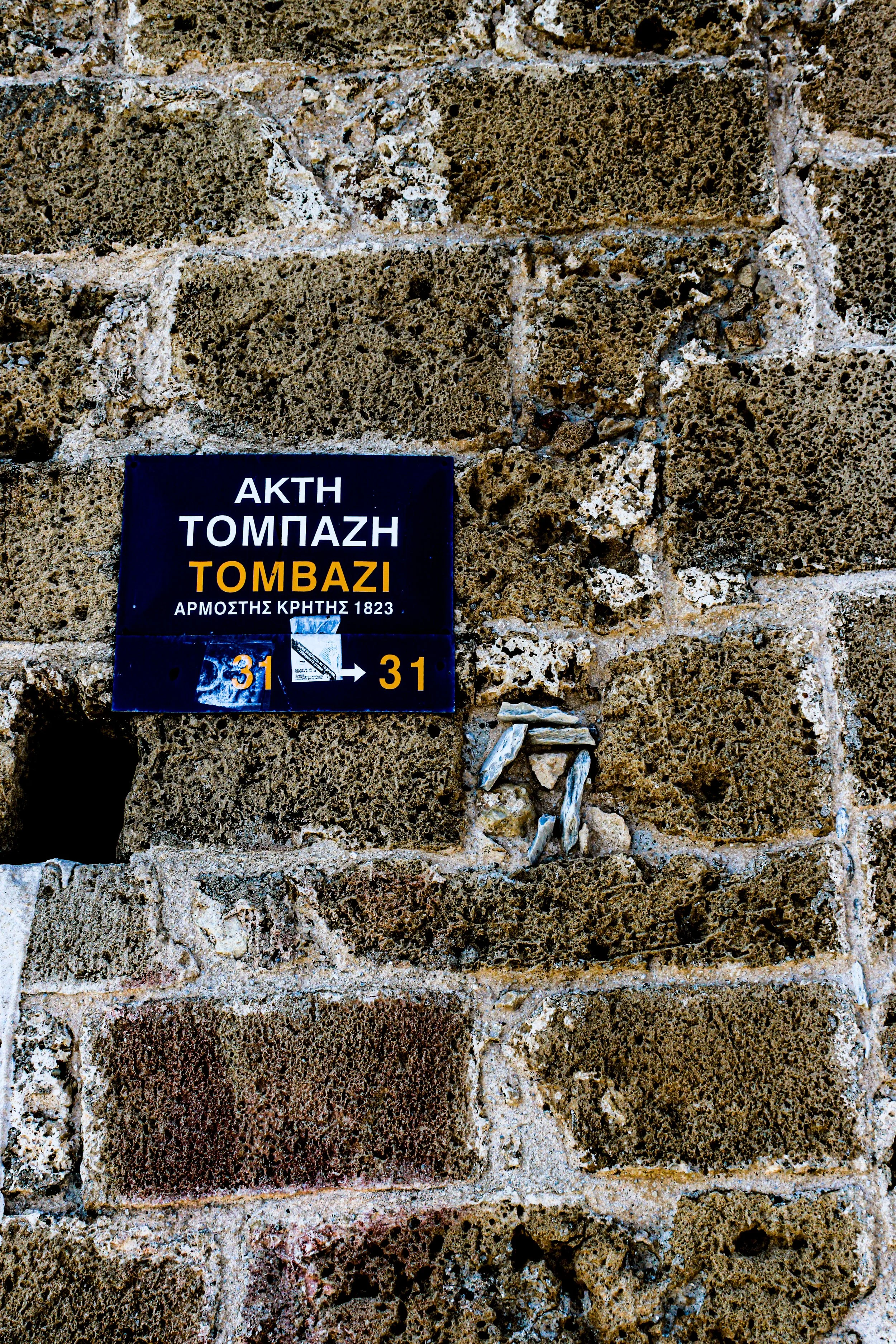 Close-up of a stone wall with rectangular stones and a small blue sign with Greek text and arrows pointing in both directions.