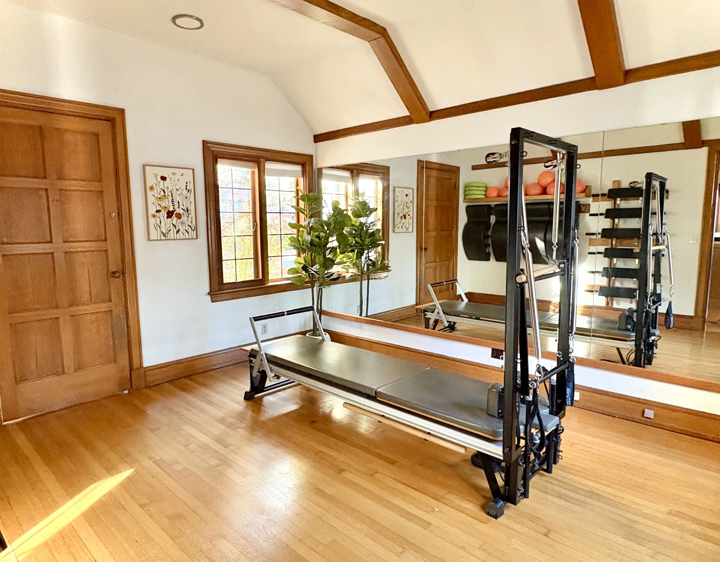 A wooden-floored home gym with large windows, a mirror, a Pilates reformer machine, and shelves with exercise balls and towels.