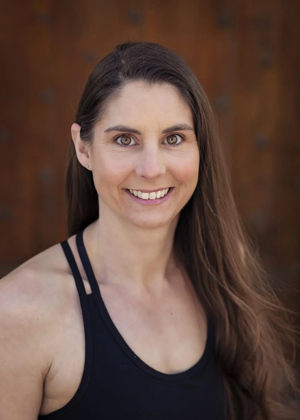 A woman with long brown hair, wearing a black tank top, smiling, with a blurred brown background.