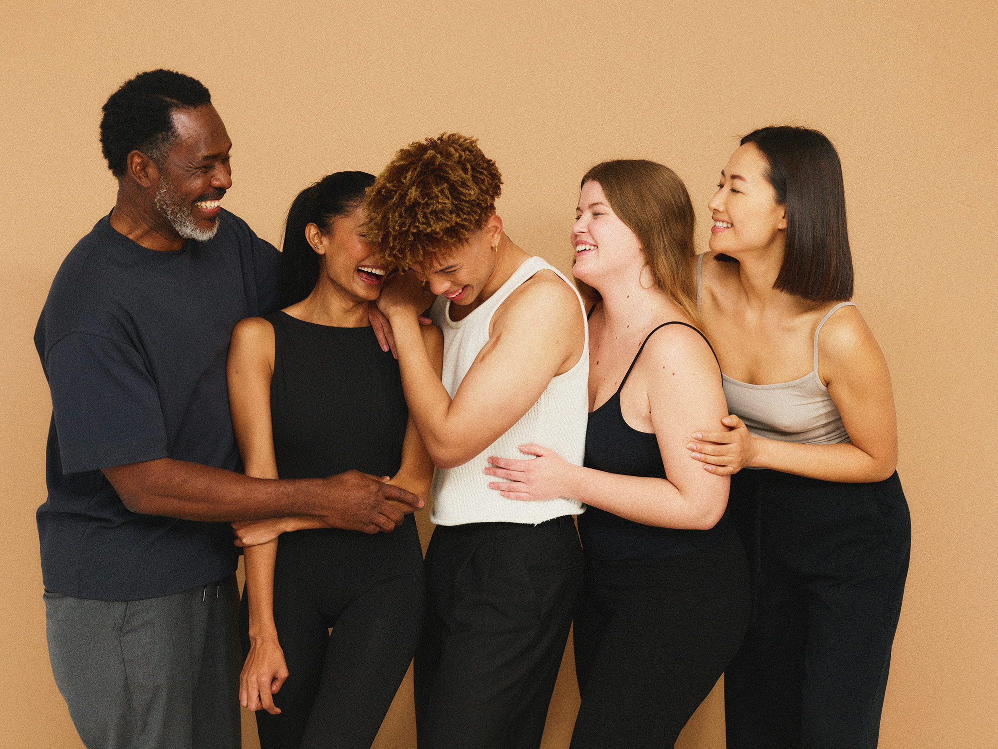 Six women and one man sharing a joyful moment, embracing and smiling against a plain beige background.