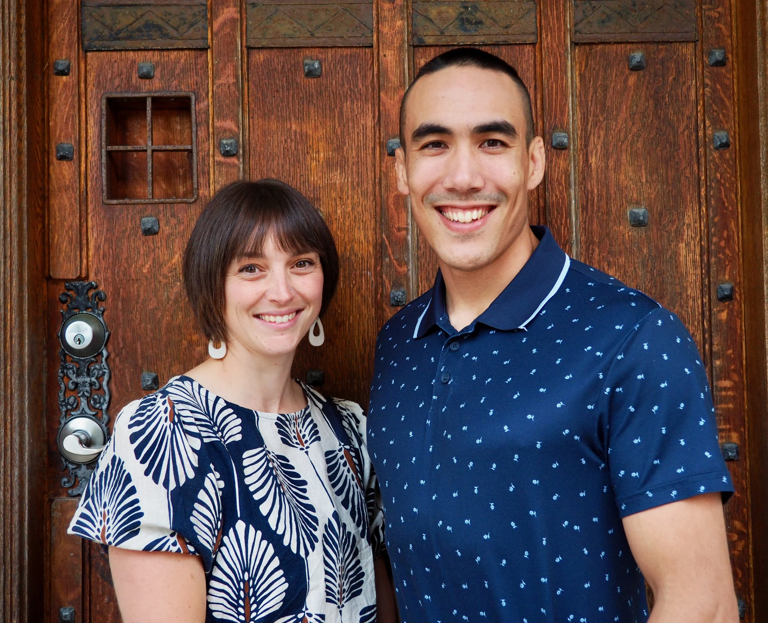 A smiling man and woman standing in front of a large wooden door with black iron details.