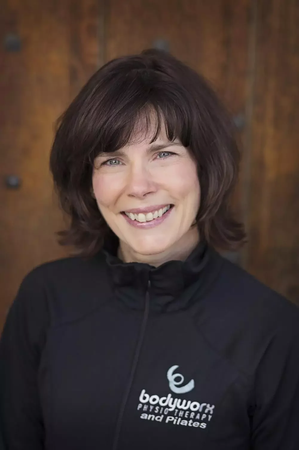 A woman with shoulder-length brown hair, smiling, wearing a black athletic jacket with 'bodywork PHYSIOTHERAPY and Pilates' logo, standing against a wooden background.