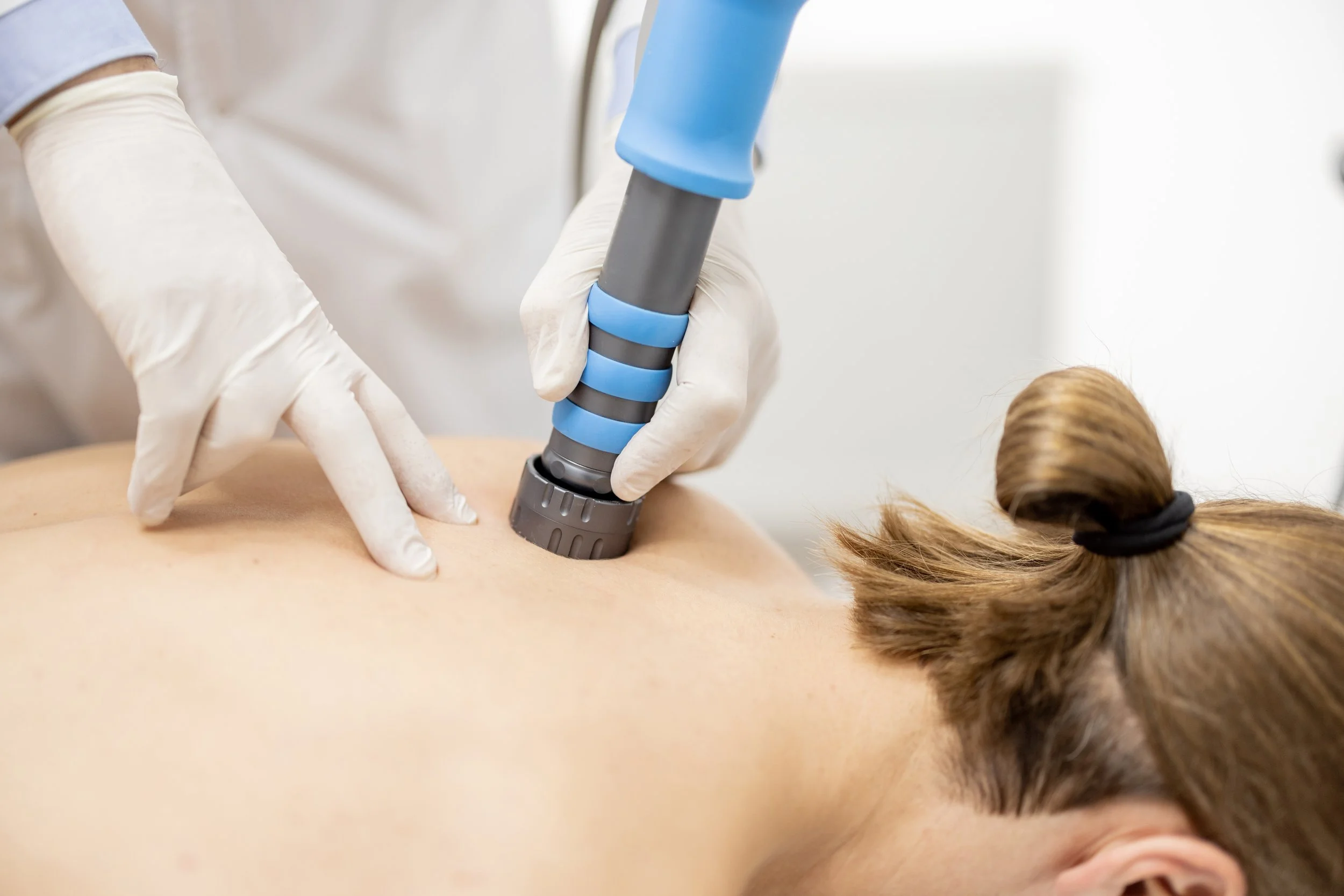 A person receiving a medical treatment with a handheld device on their back while lying face down, with a healthcare worker wearing gloves.