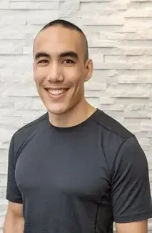 Young man with short hair, smiling, wearing a dark gray t-shirt, standing in front of a white brick wall.