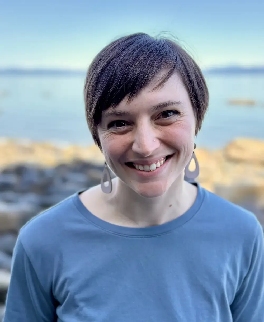 A woman with short dark hair, smiling, wearing a blue shirt and earrings, with a blurred body of water and rocks in the background.