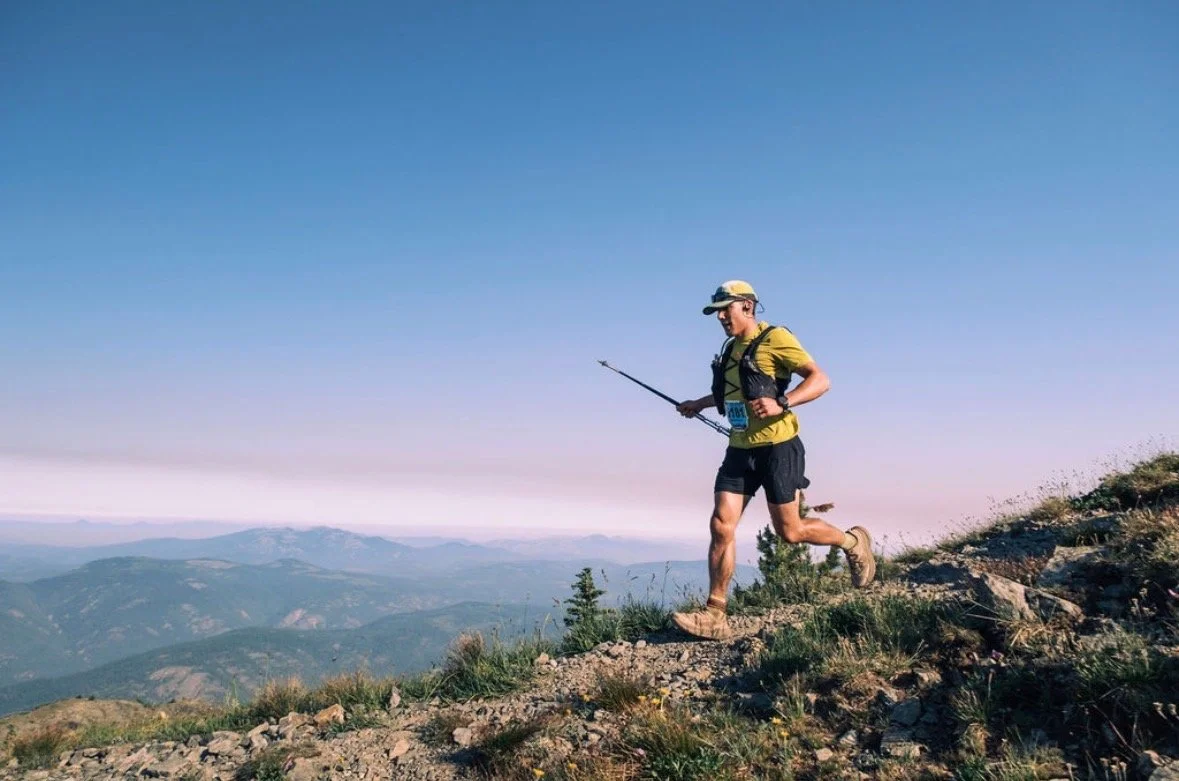 A person trail running on a mountain trail with a scenic mountain range in the background under a clear blue sky.