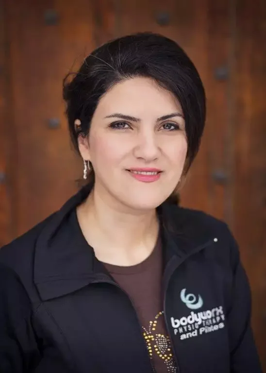 A woman with short dark hair wearing a black jacket with a logo that says 'bodywork physical therapy and Pilates' and a brown shirt underneath, standing in front of a wooden wall.