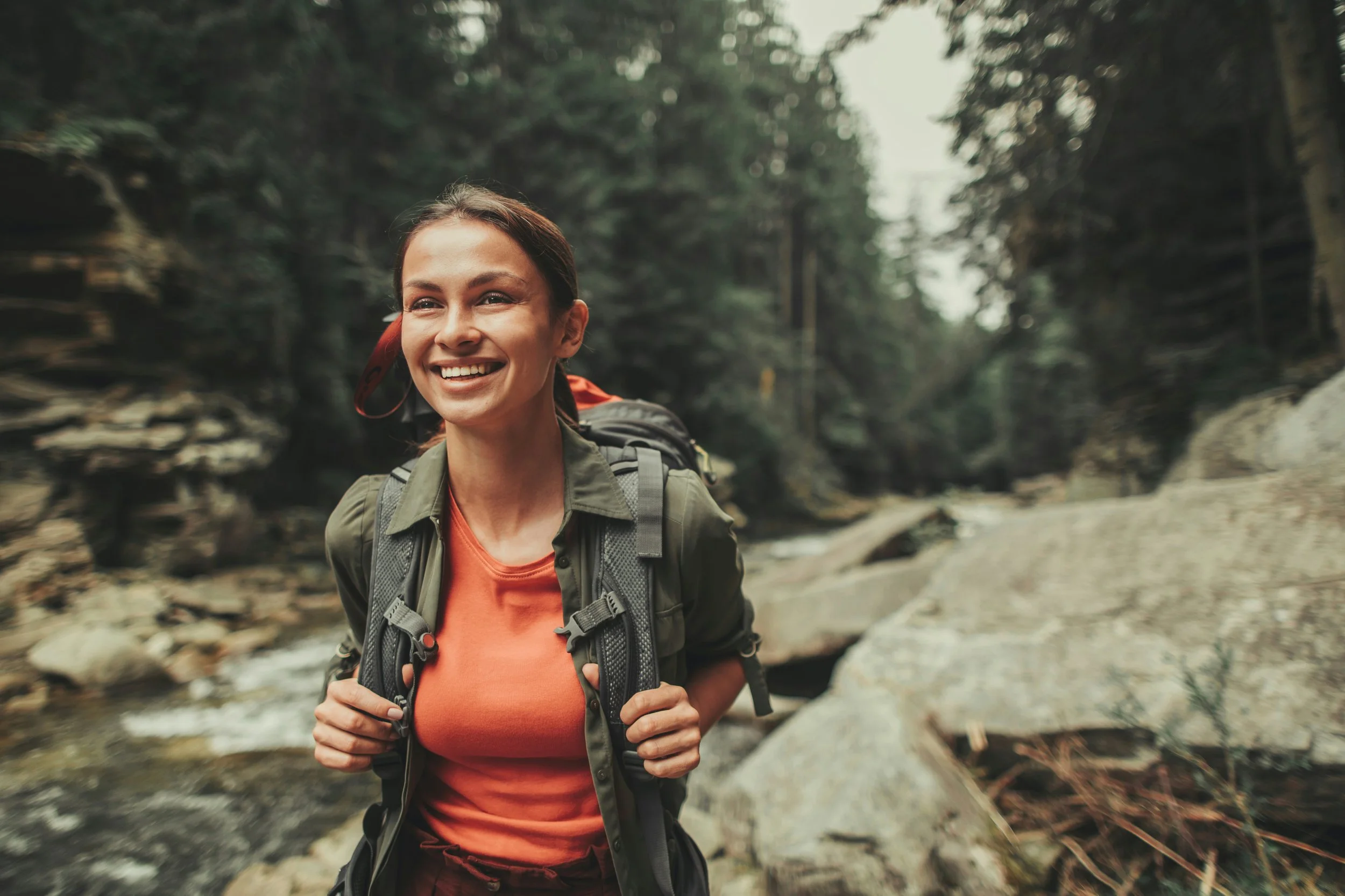 A smiling woman with long brown hair, wearing an orange shirt and a green jacket, carrying a backpack, hiking in a forest with a stream and trees in the background.