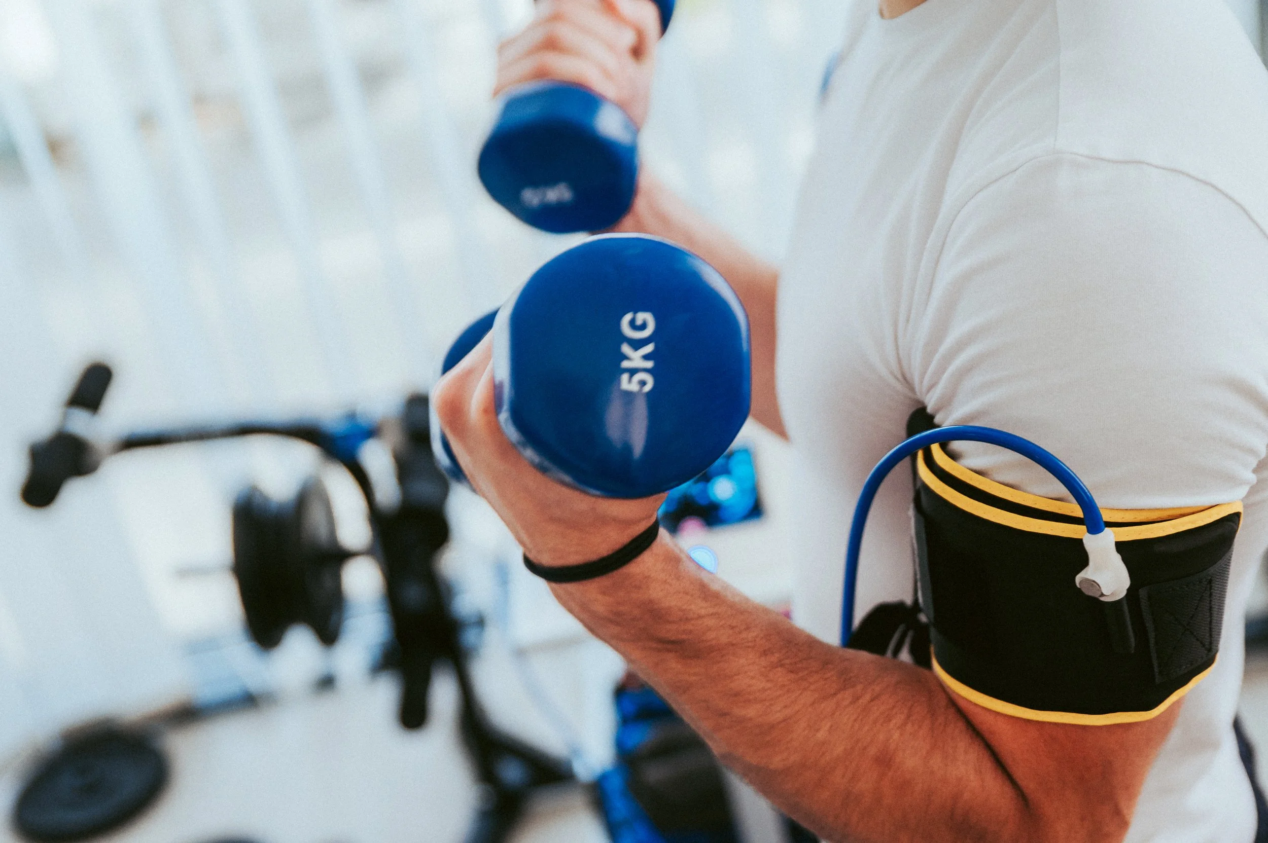 A person with a tattooed arm lifting a blue dumbbell labeled '5 KG' in a gym. The person is wearing a white and black shirt and a black and yellow fitness tracker or armband on their bicep. Several pieces of gym equipment are visible in the background.