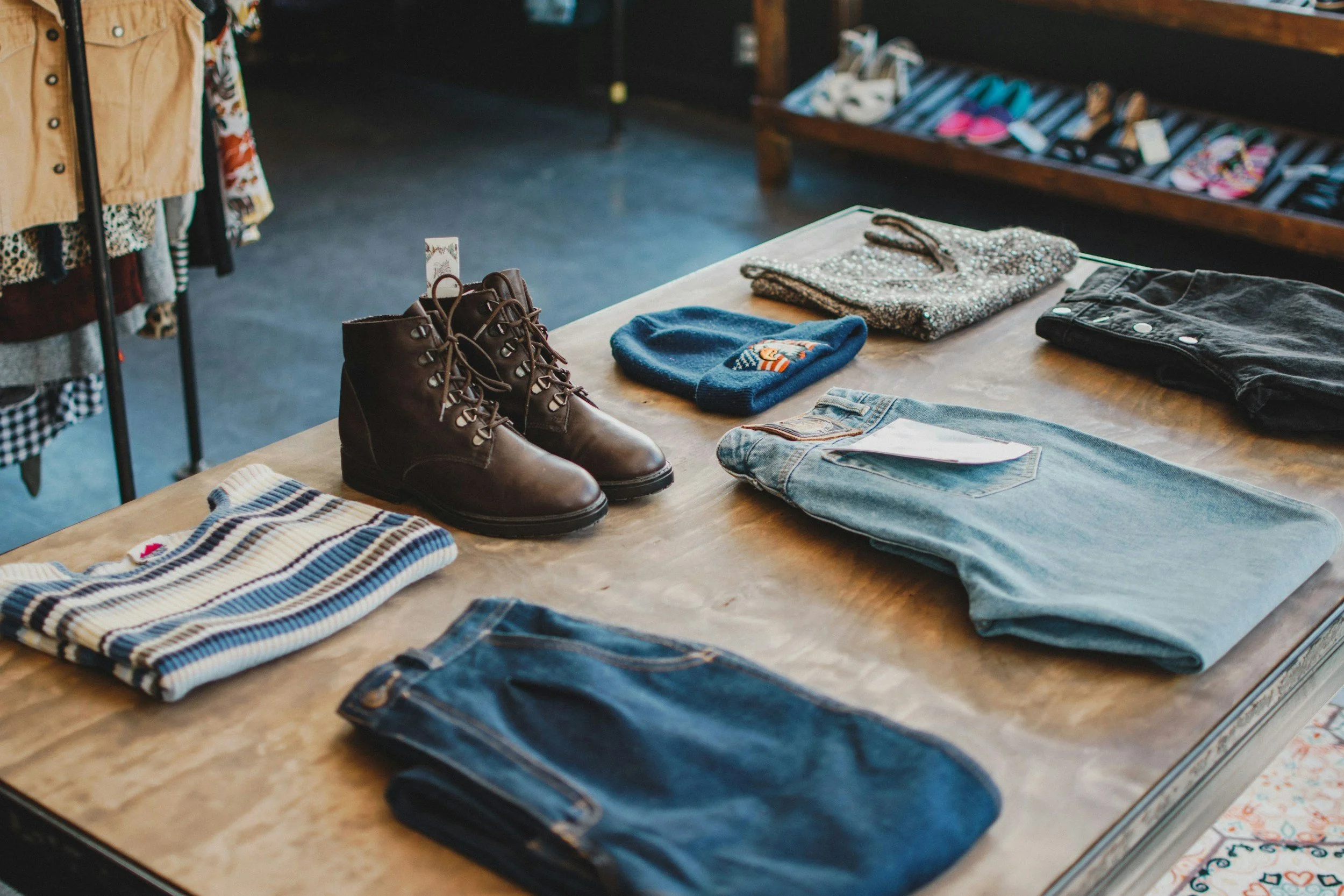 A wooden table displays folded clothing items including jeans, sweaters, and a striped shirt, with a pair of brown combat boots nearby, inside a retail store.