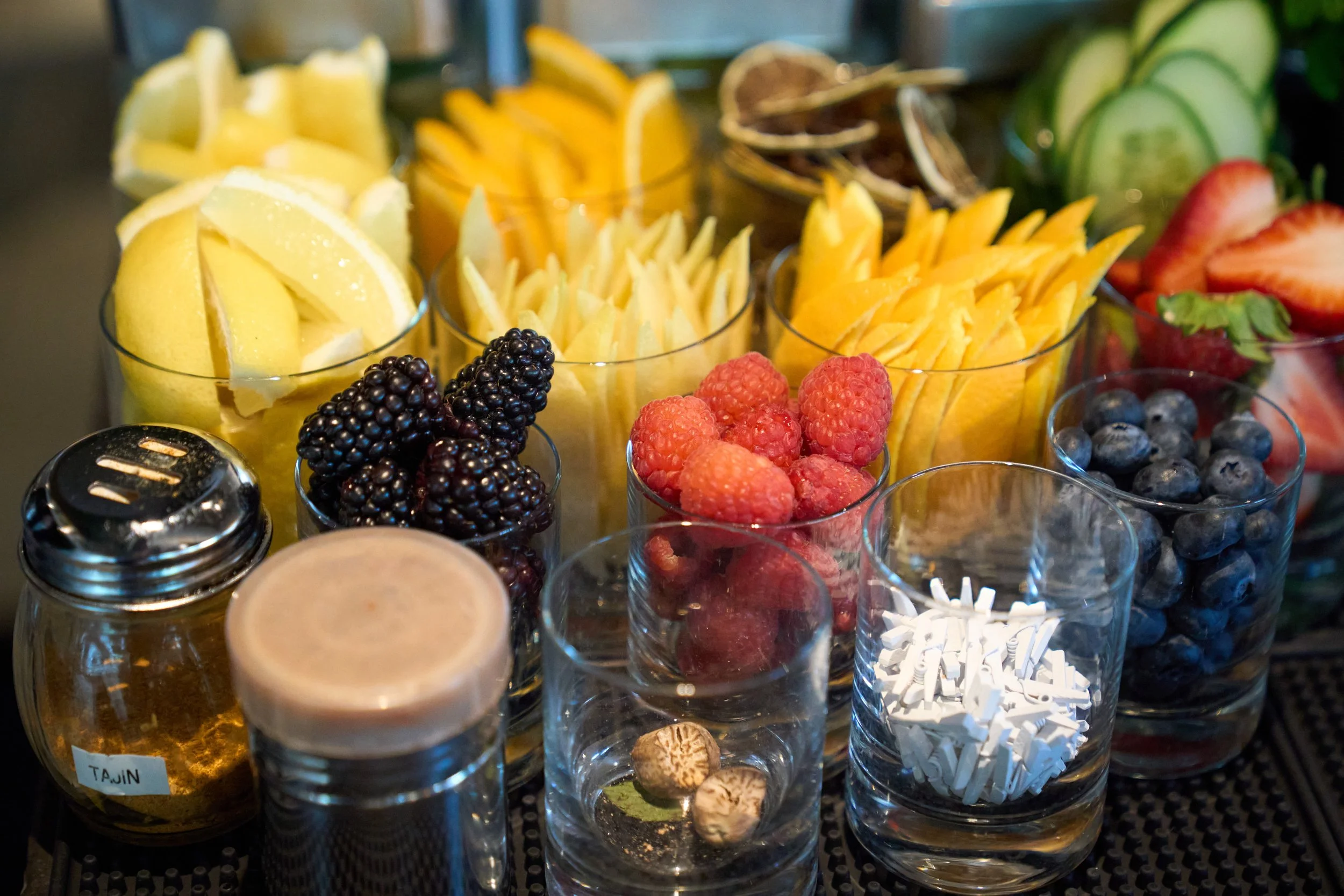 An assortment of sliced and whole fresh fruits in glass containers, including lemons, pineapple, blackberries, raspberries, strawberries, blueberries, and pepper to the right. In the foreground, small jars contain honey and a creamy sauce, with shred