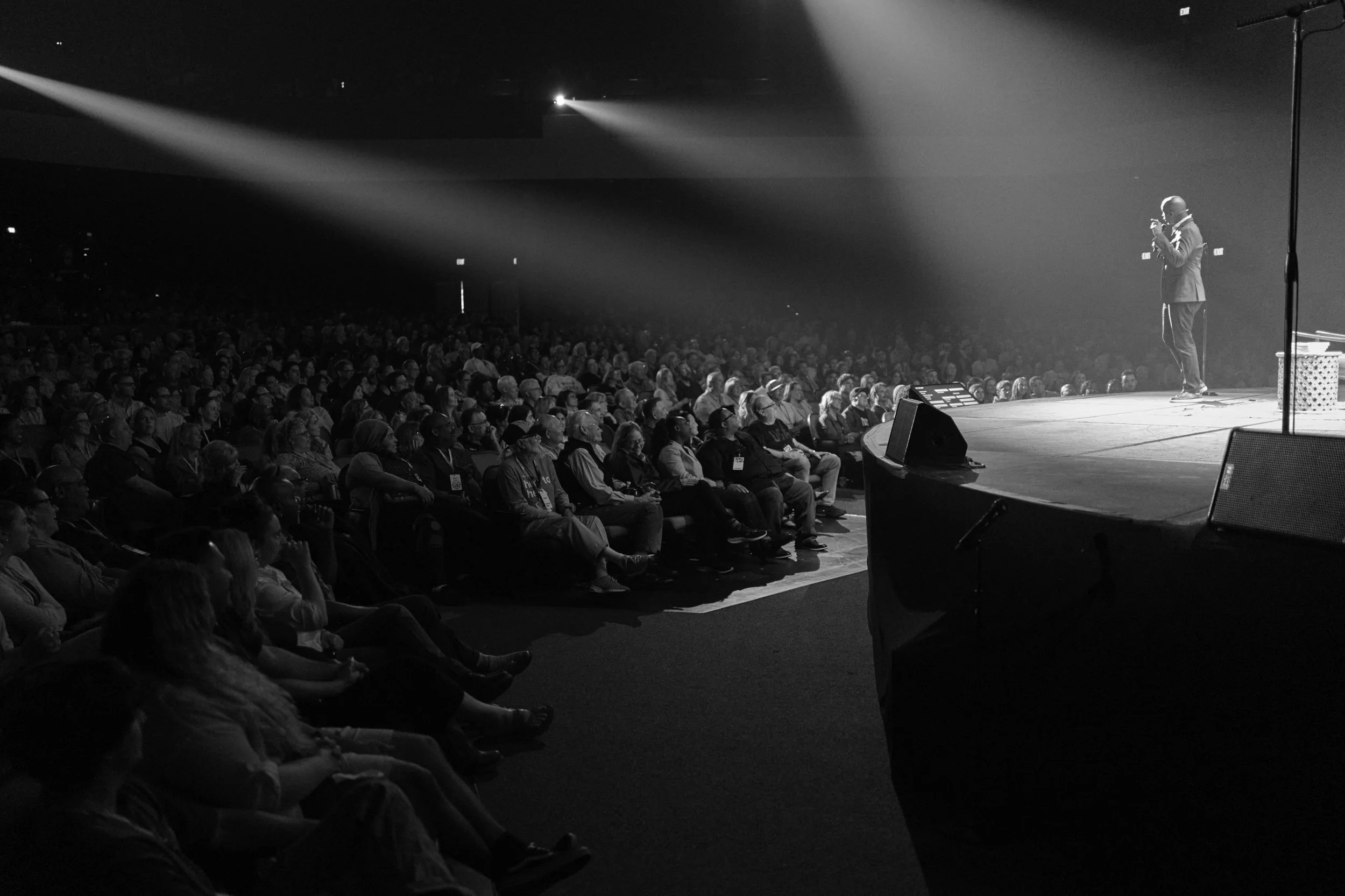 Black and white image of a speaker on stage holding a microphone, addressing a large audience in an auditorium, with stage lights focused on speaker and audience members seated and watching attentively.