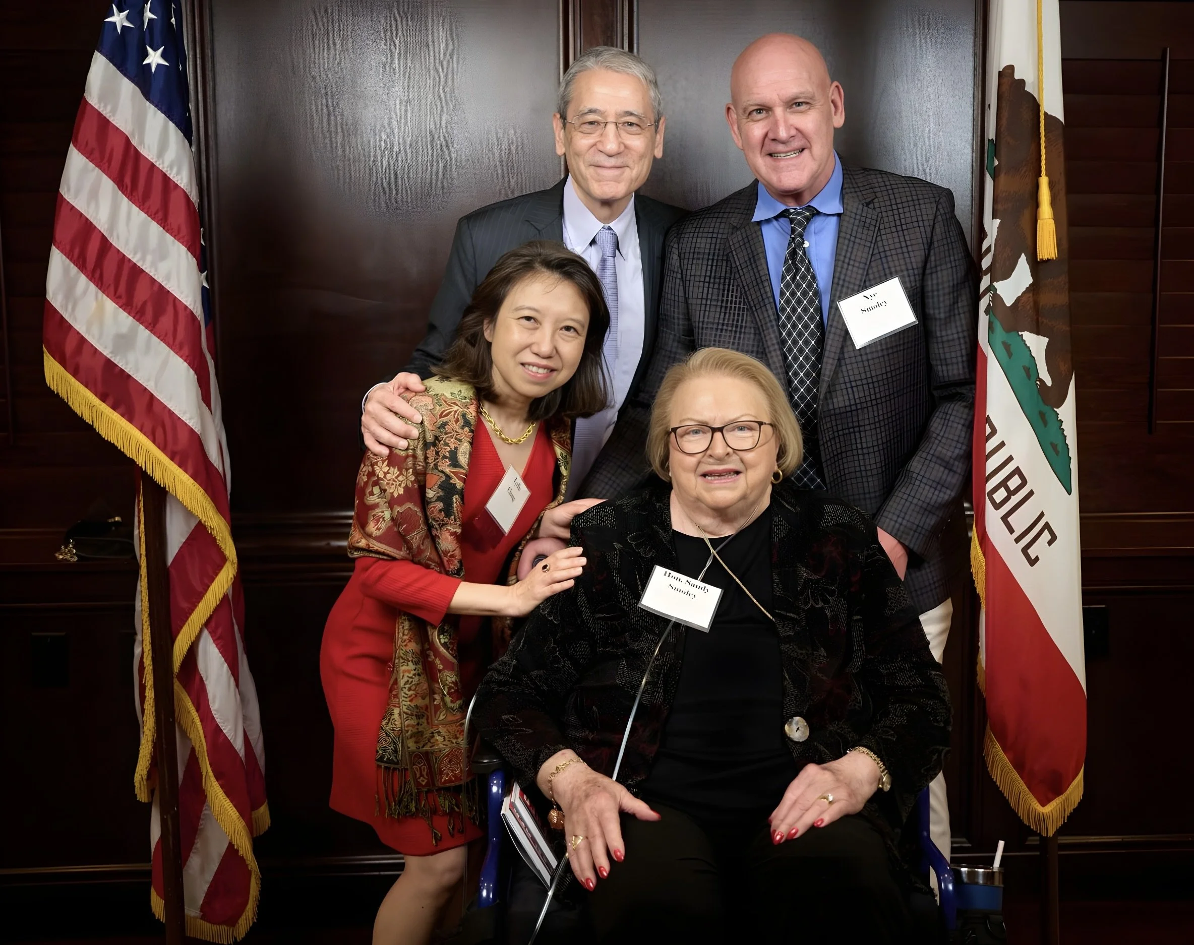 A group of five diverse, smiling individuals gathered together indoors in front of two flags, the United States flag and the California state flag, with a dark wood-paneled wall in the background.