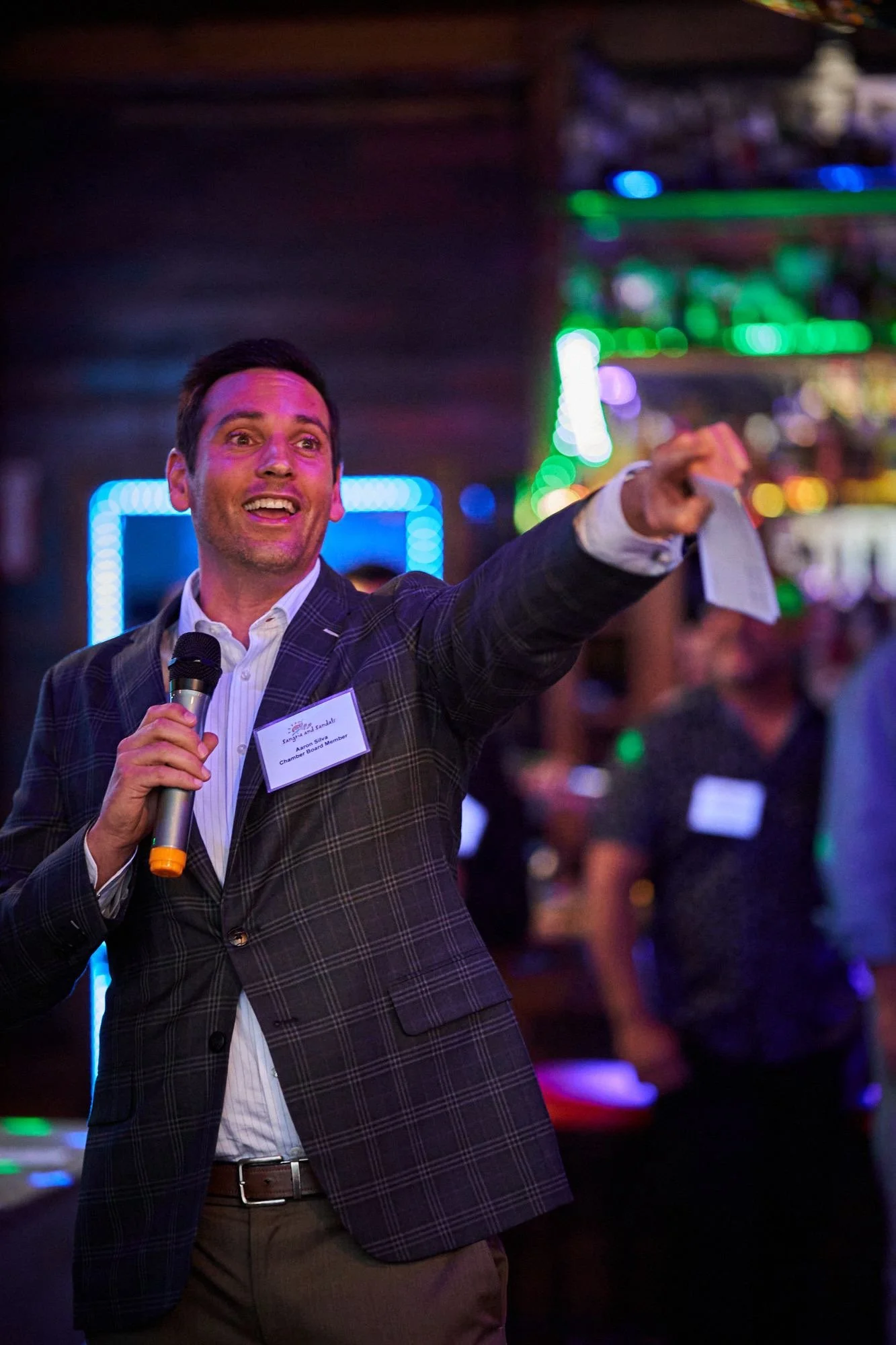 A man in a plaid blazer and dress shirt, holding a microphone, is pointing and speaking passionately at an indoor event with colorful neon lights.