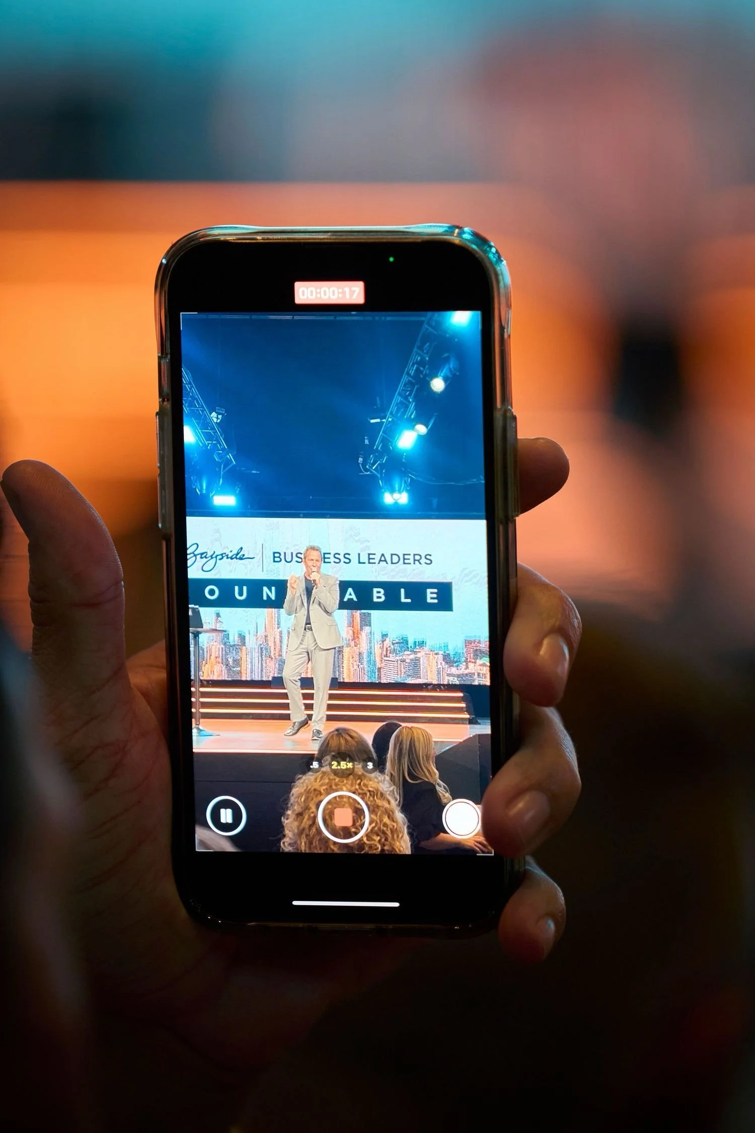A person holding a smartphone capturing a speaker on stage at a conference or seminar with the words "BUSINESS LEADERS" and "UNAVAILABLE" displayed on a screen behind him.