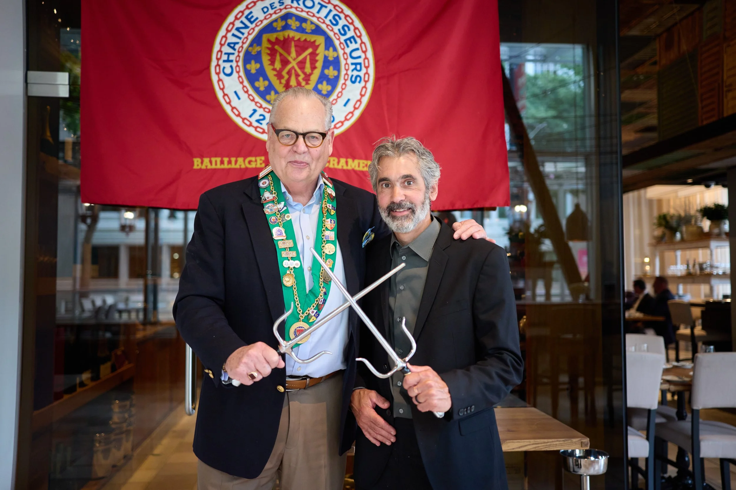 Two men standing together at an event in front of a red flag with an emblem of the Scouts of France. The man on the left wears glasses, a suit, and a colorful sash with medals. The man on the right has gray hair and a beard, and wears a dark suit wit