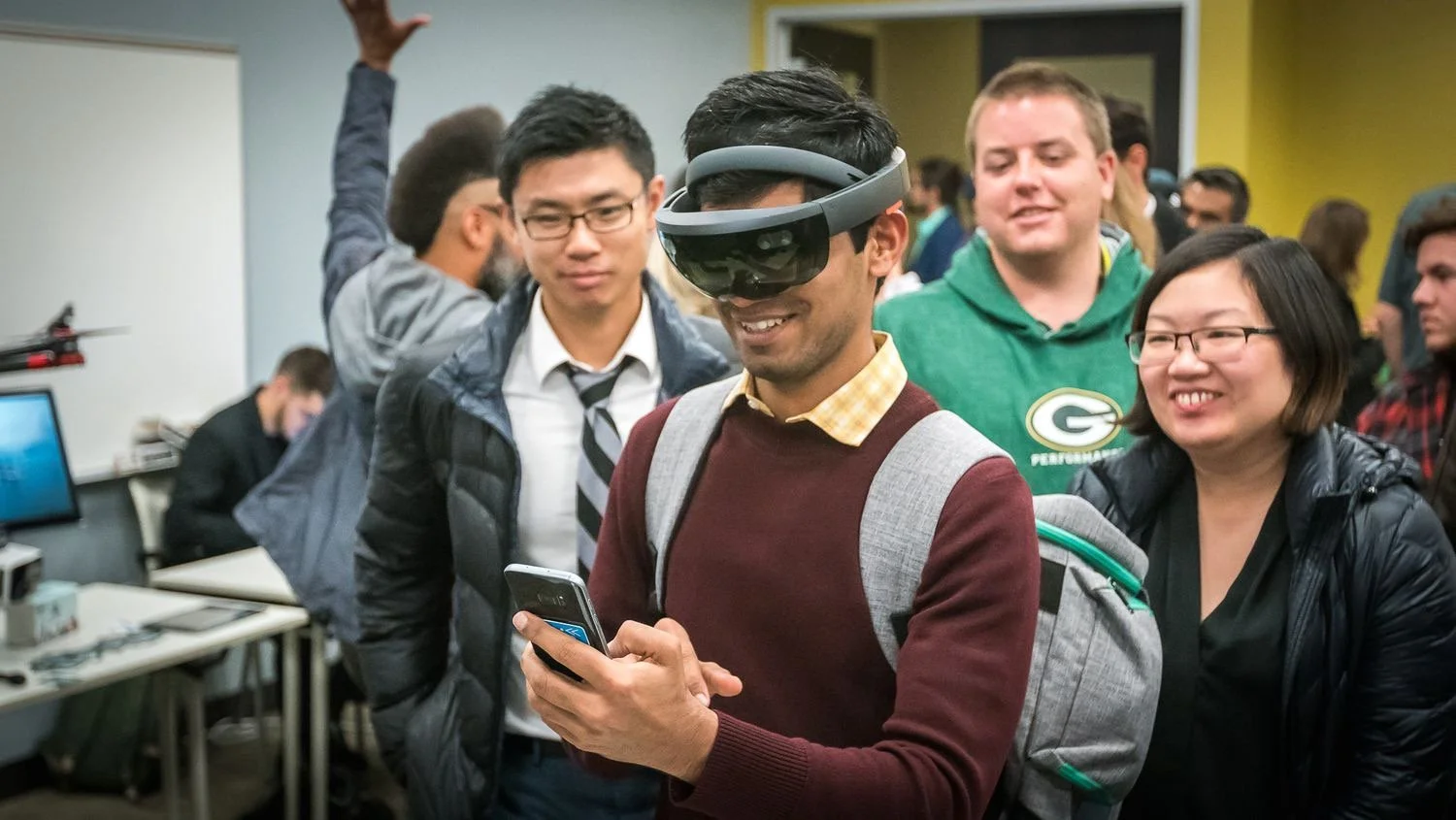 A man wearing a virtual reality headset and a backpack while smiling and looking at his phone, surrounded by smiling people, in an indoor tech event or classroom.