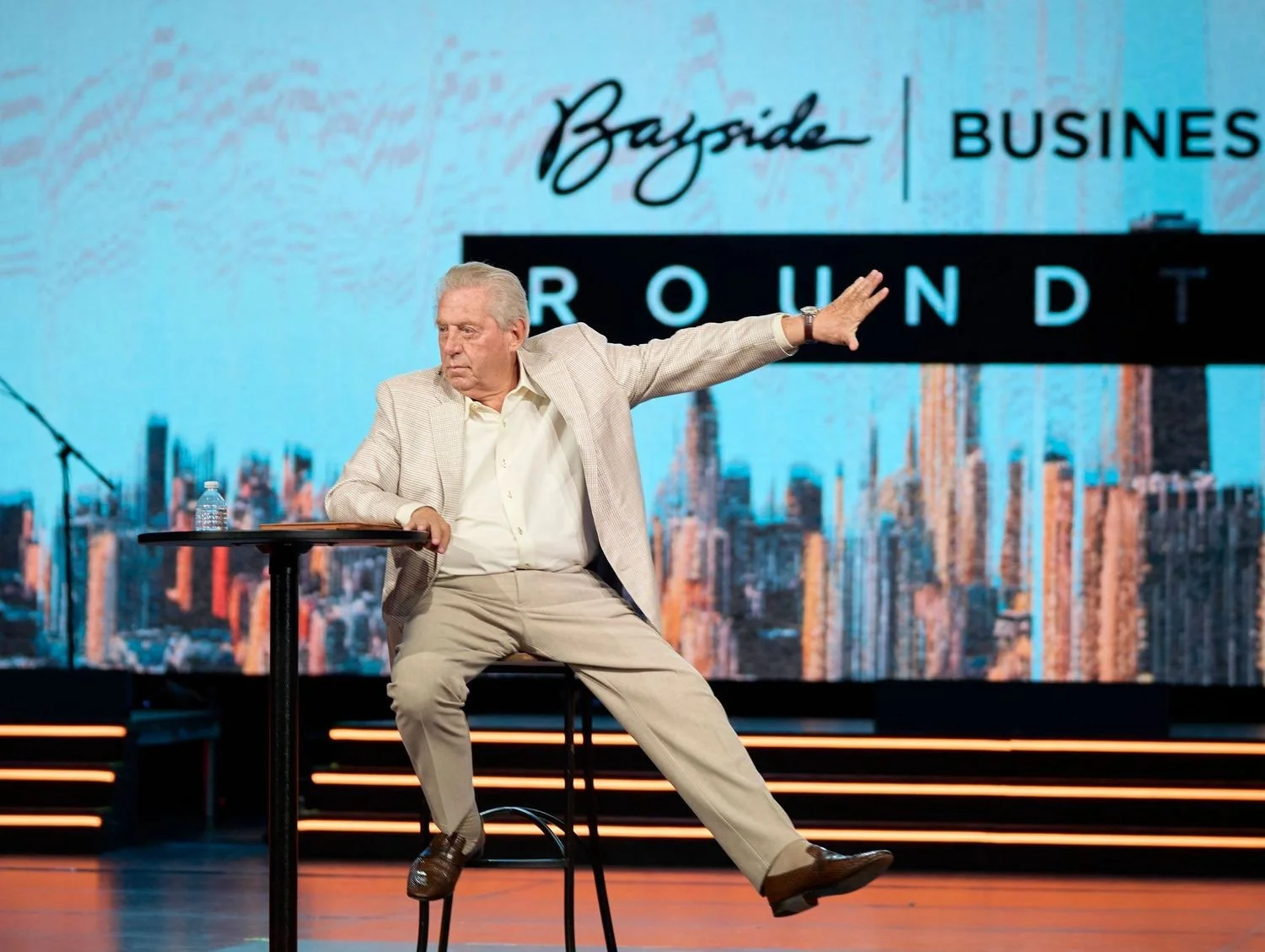 An elderly man in a beige suit and white shirt, sitting on a tall chair on a stage, gesturing with one arm extended to the right, with a water bottle on a small table beside him. Behind him is a large screen displaying the words 'Bay Area Business Ro