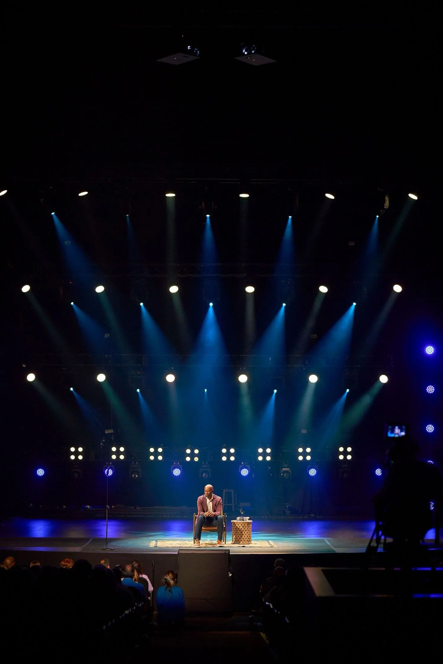 Stage with a performer sitting on a chair at center, surrounded by stage lighting and an audience in the foreground.