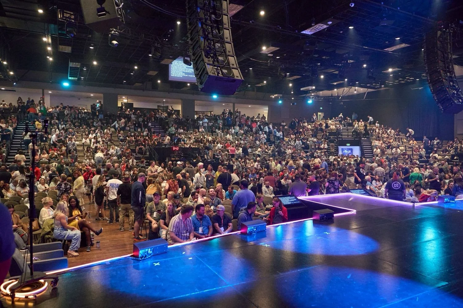A large auditorium filled with seated and standing people, with a stage in the foreground lit with colorful lights, and an overhead sound system with large speakers.
