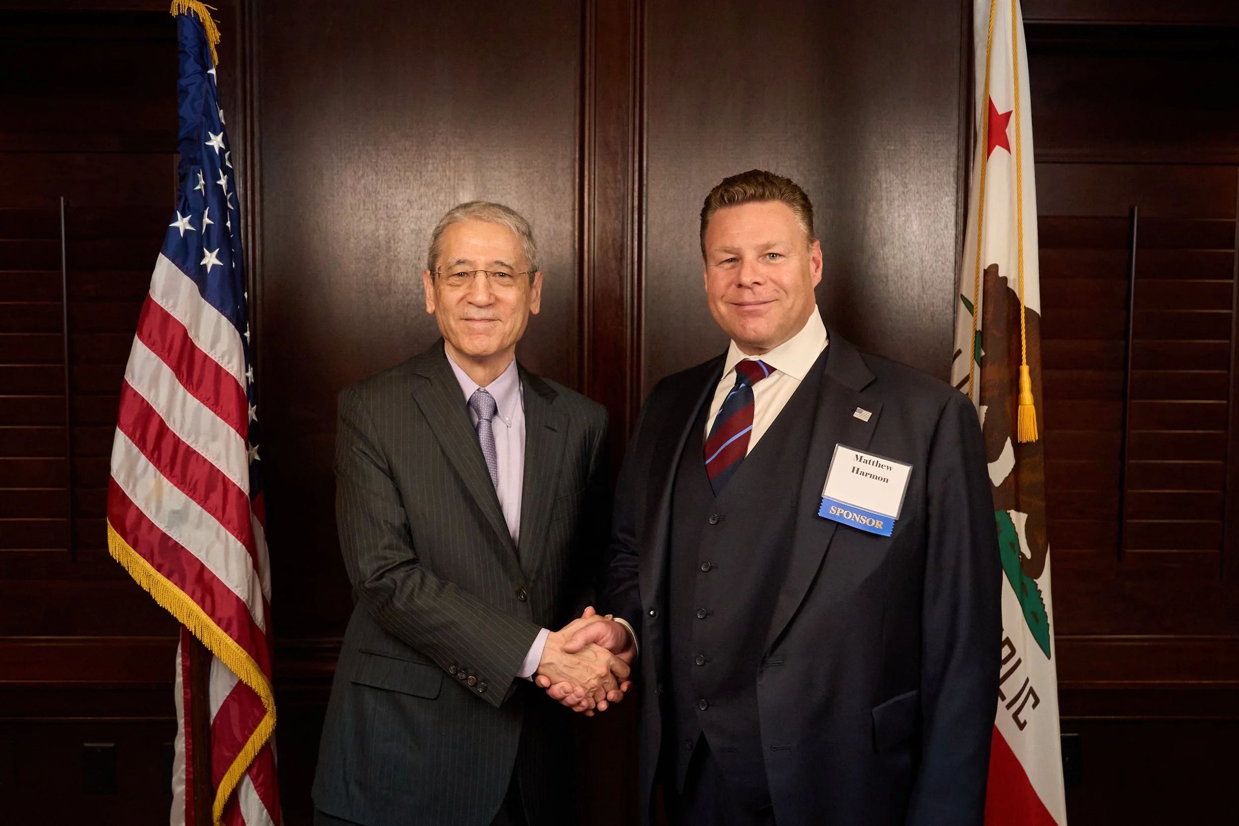 Two men in suits shaking hands and smiling, standing between American and California flags in a wood-paneled room, with each man wearing a name badge.