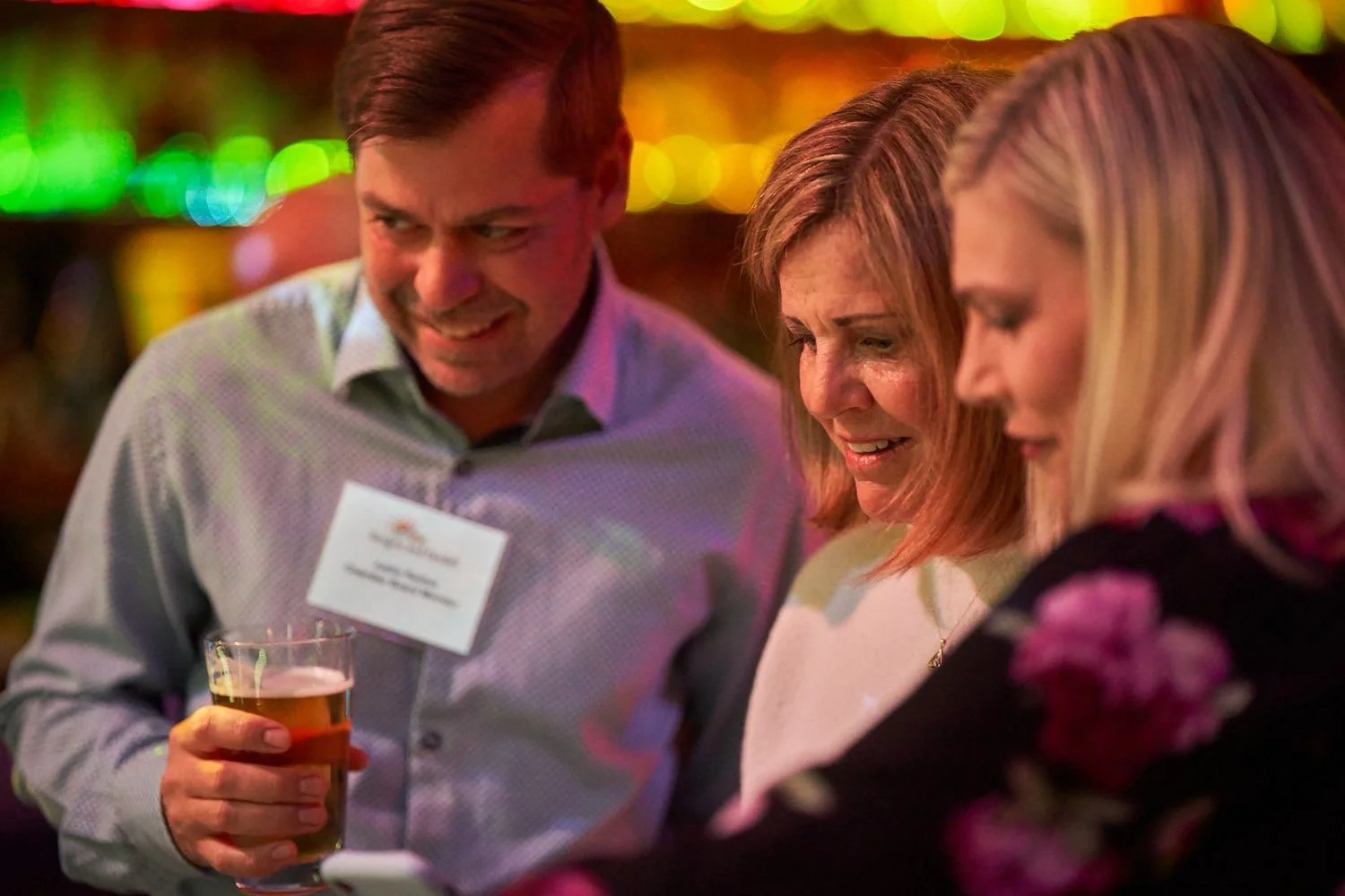 Three people at a social event, two women and one man, looking at a phone together in a dimly lit setting with colorful lights in the background. The man holds a drink in his hand.